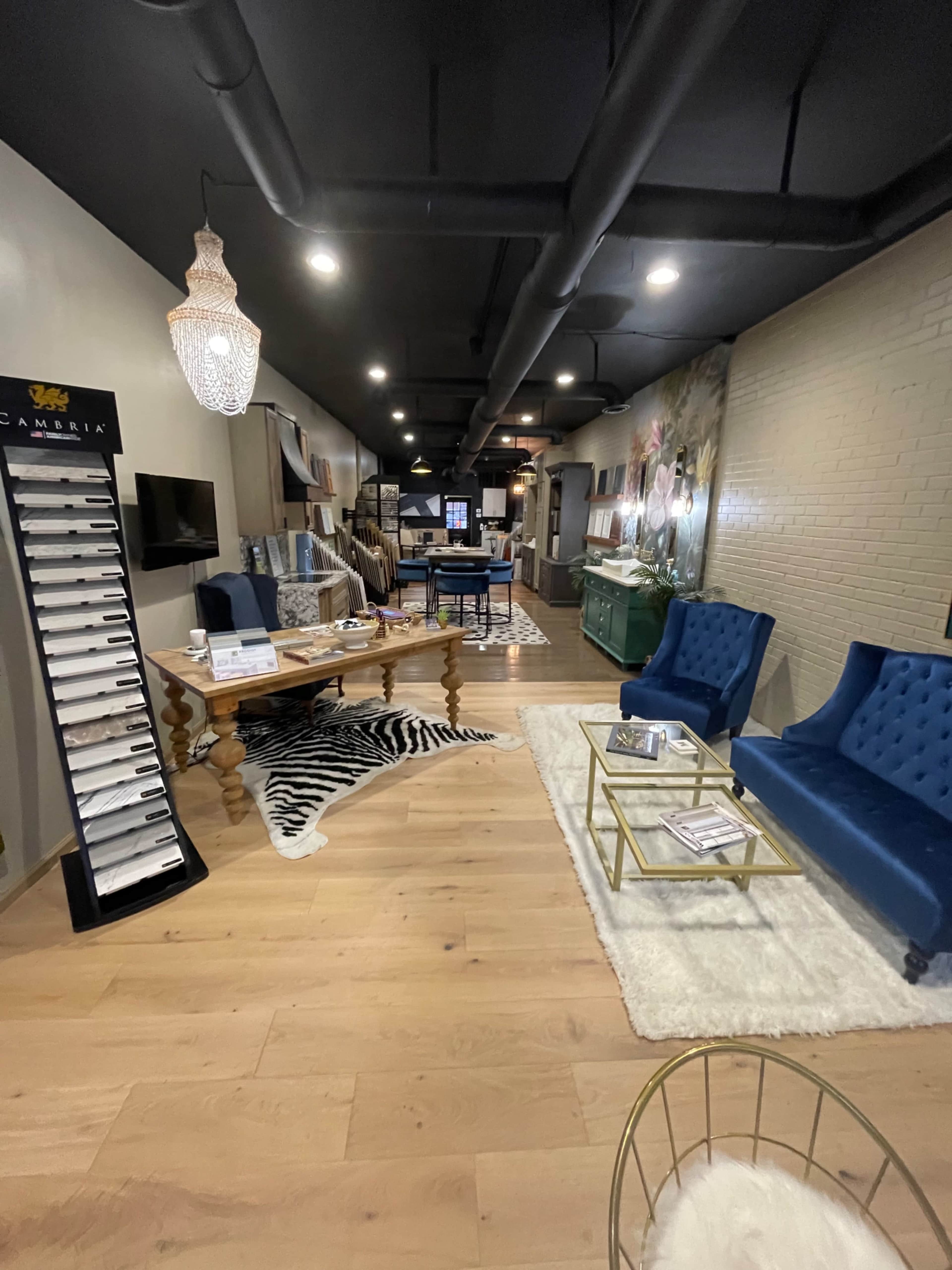 The image shows a modern interior space featuring blue upholstered chairs, a wooden table, and a display stand along with decorative elements like rugs and wall art.