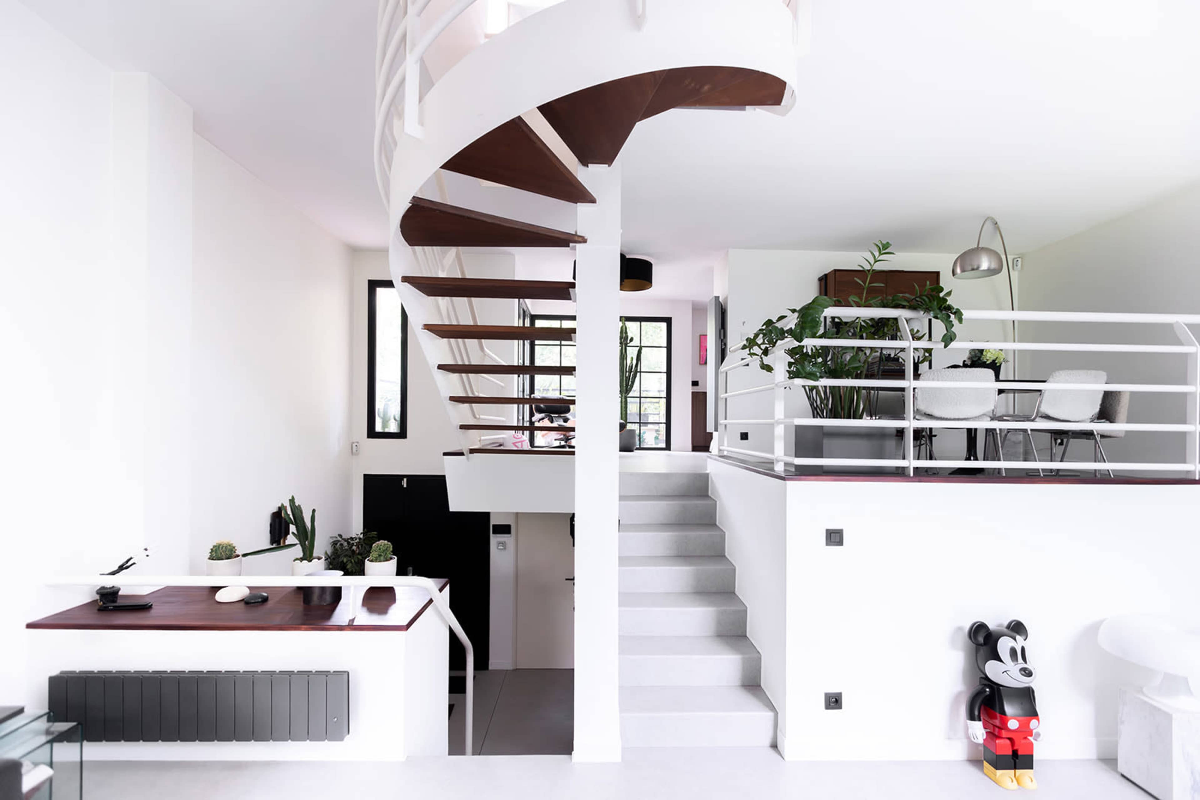 A modern interior with a spiral staircase, a dining area with plants, and artistic decor, including a figure resembling a popular cartoon character.