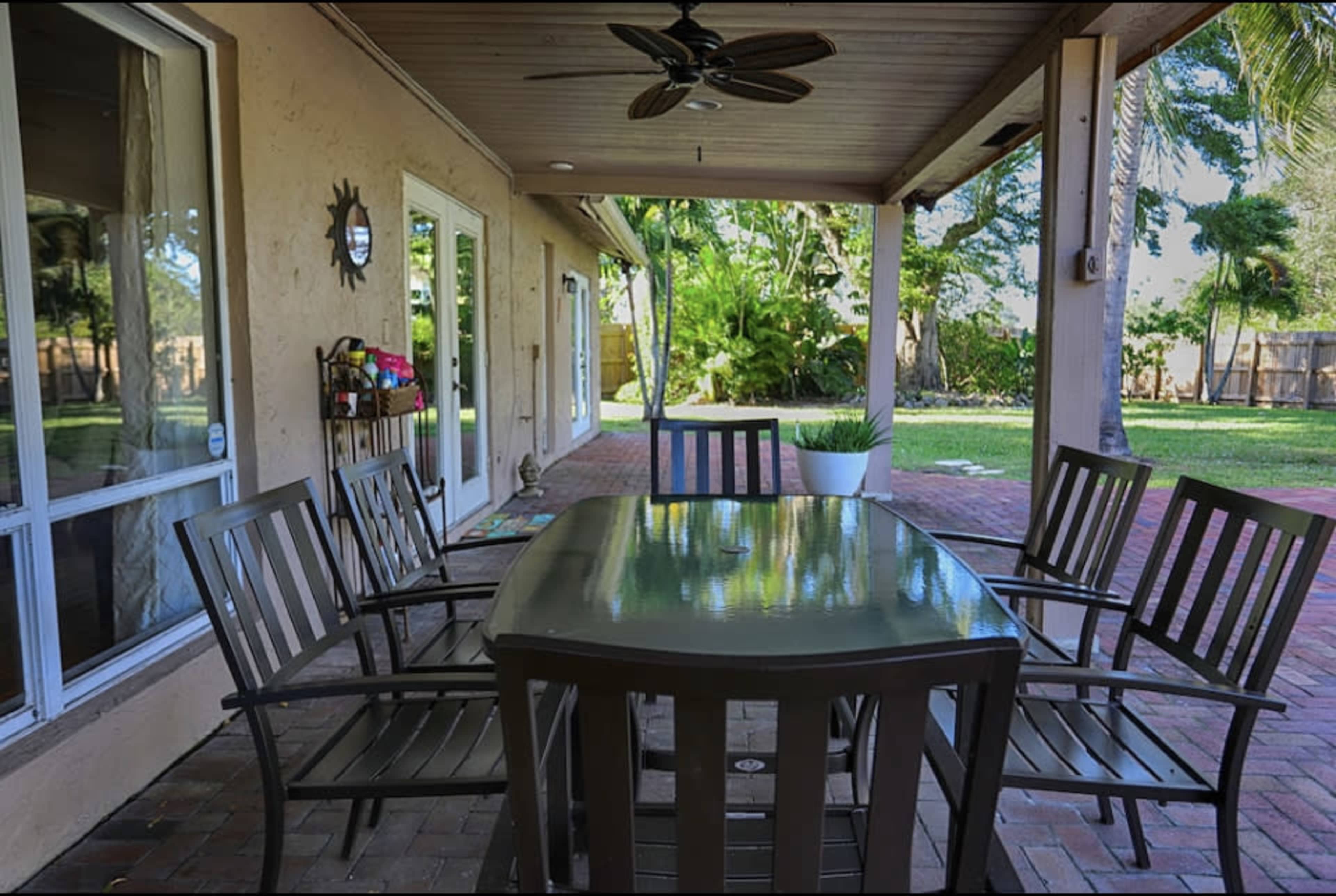 A patio area features a rectangular table surrounded by six metal chairs, beneath a ceiling fan, with a view of a backyard and scattered greenery.