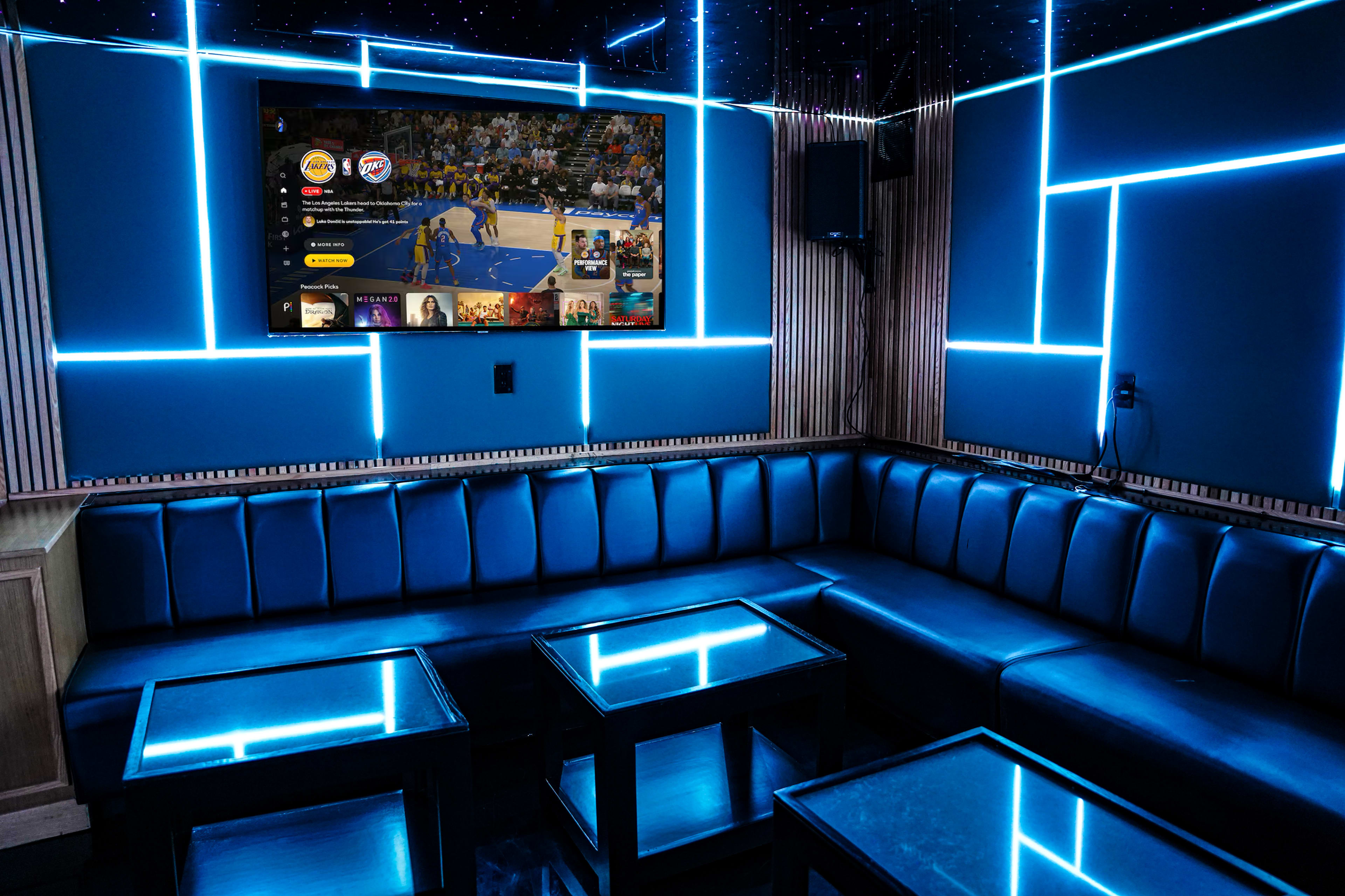 A modern entertainment lounge features a dark blue seating area with illuminated geometric wall designs and a large TV displaying a basketball game.