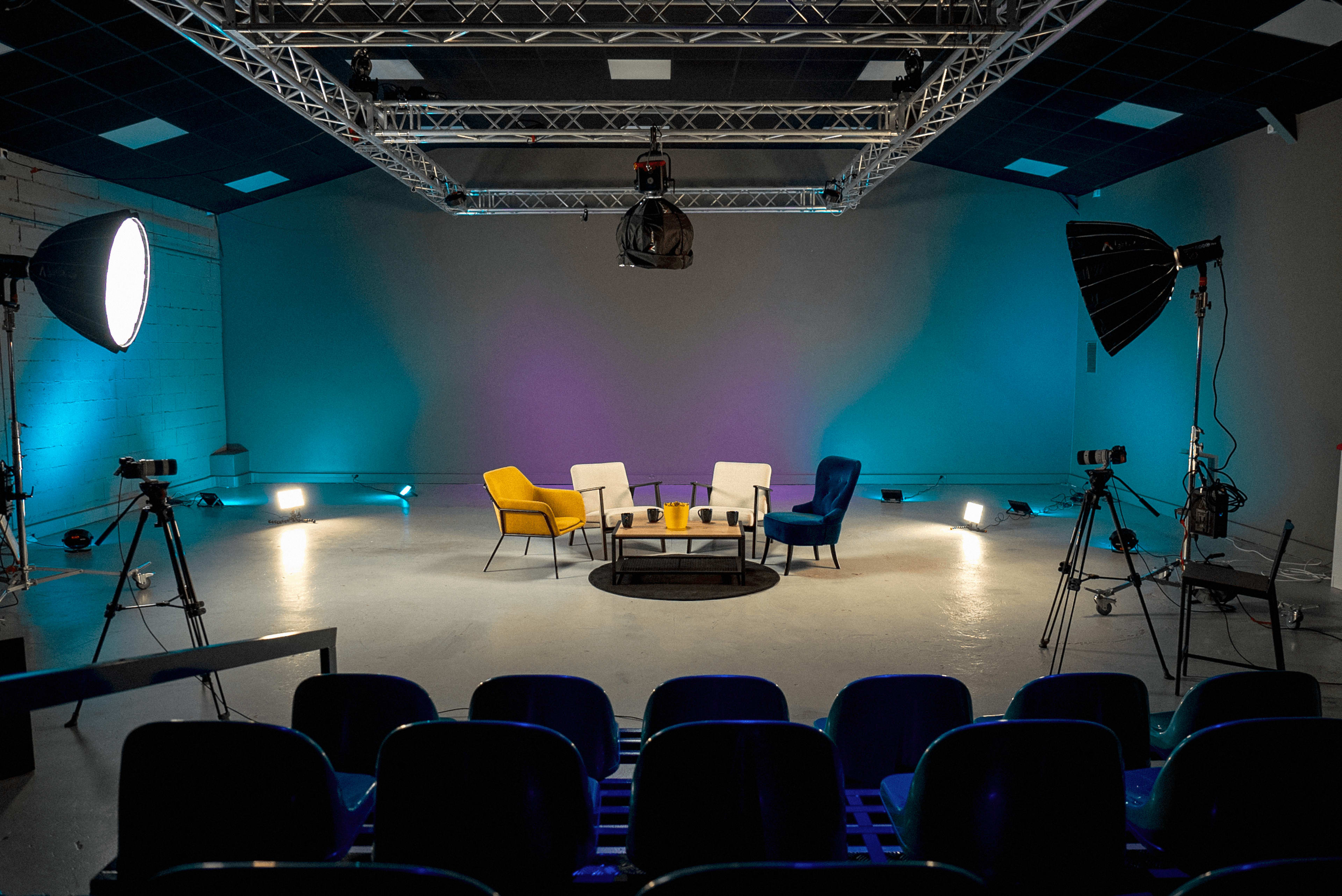 A modern studio setup features a circular seating arrangement with colored chairs, flanked by lighting equipment and cameras, against a neutral backdrop.