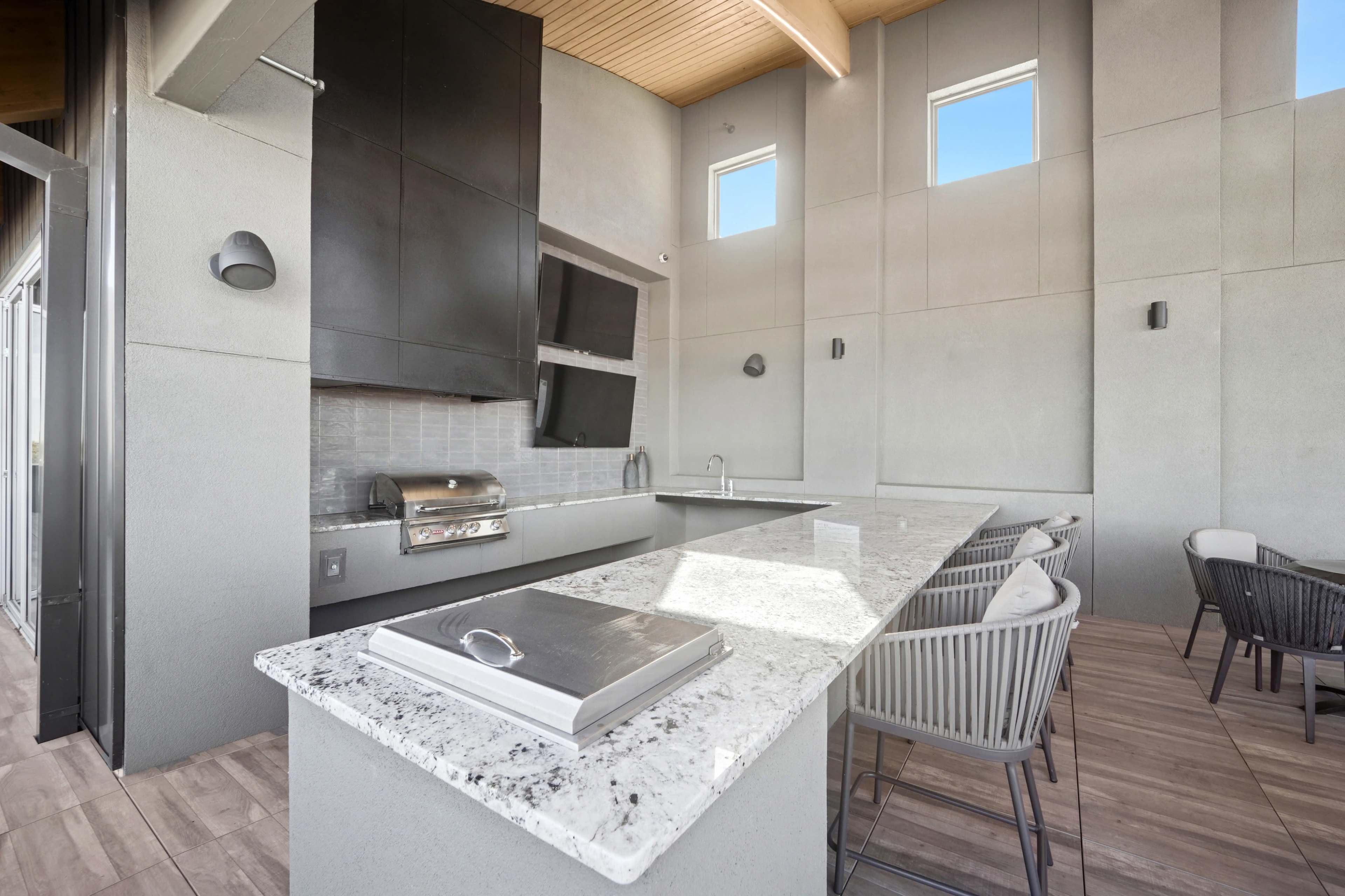 The image shows a modern outdoor kitchen area with a granite countertop, built-in grill, and bar seating.