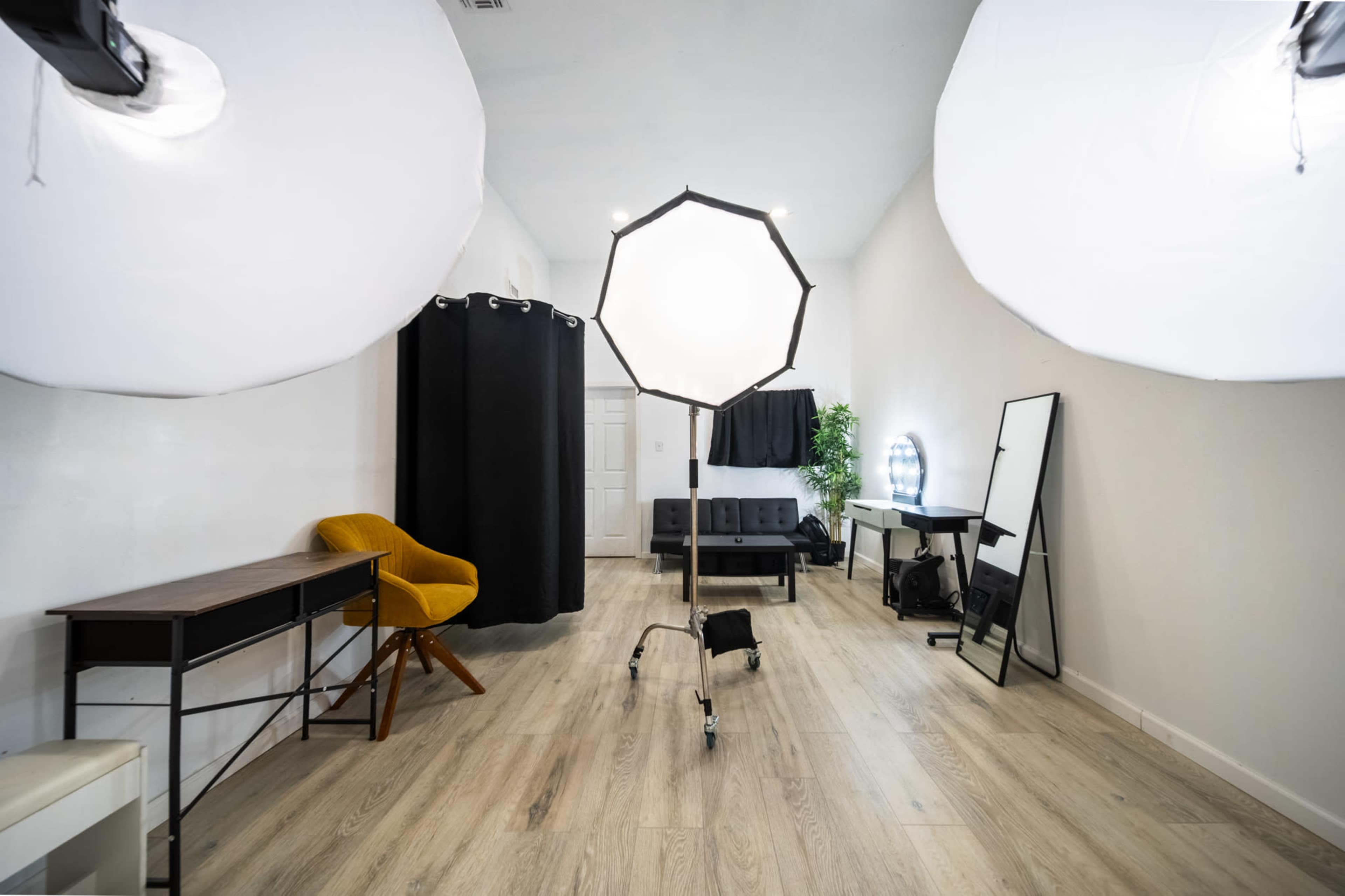 The image shows a well-lit studio space featuring photography equipment, a mirror, a yellow chair, and a black couch against a plain white wall.