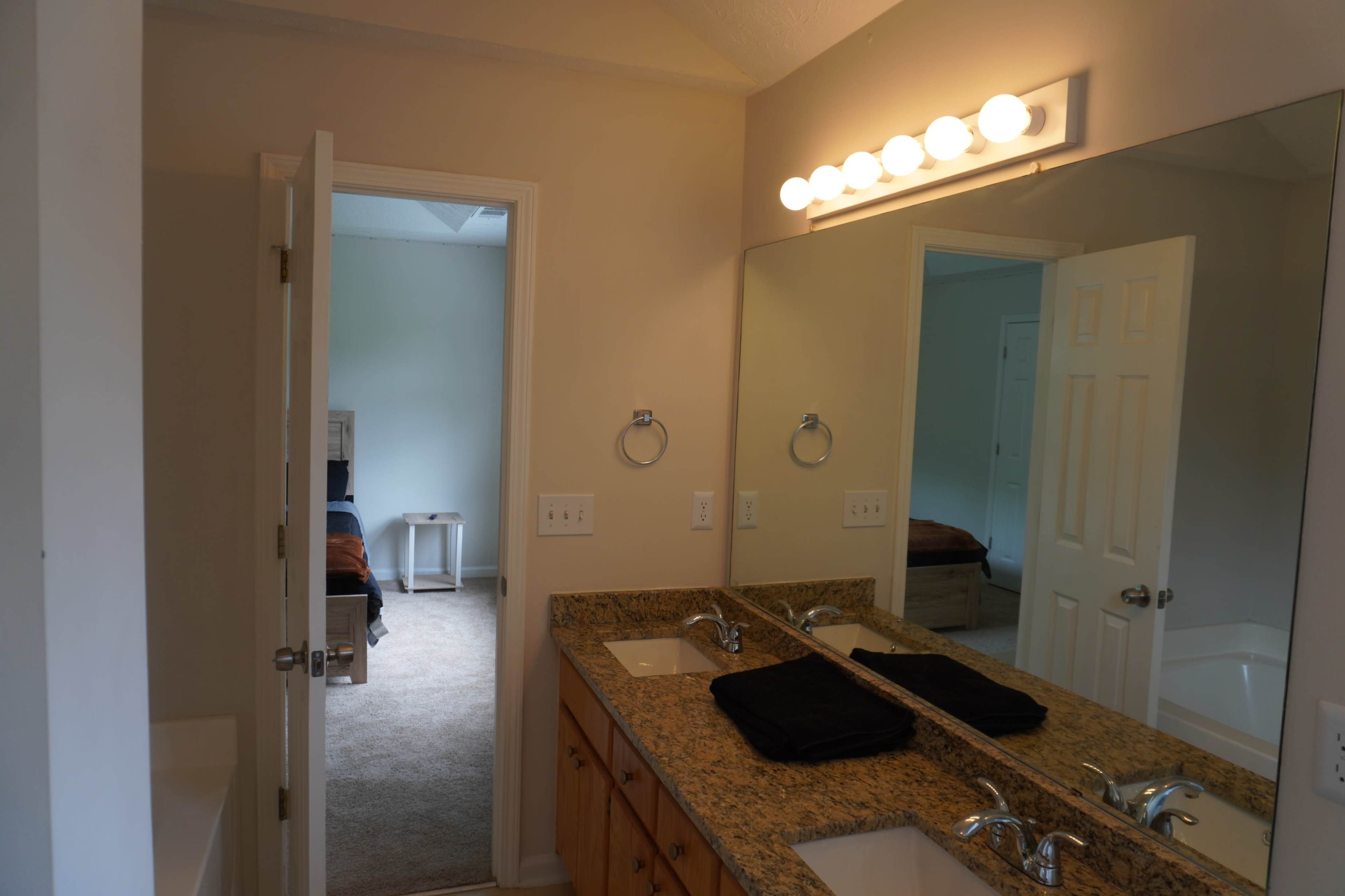 A bathroom features a double vanity with a granite countertop, illuminated by a row of light bulbs, and mirrors reflecting an adjacent room with carpeted flooring.