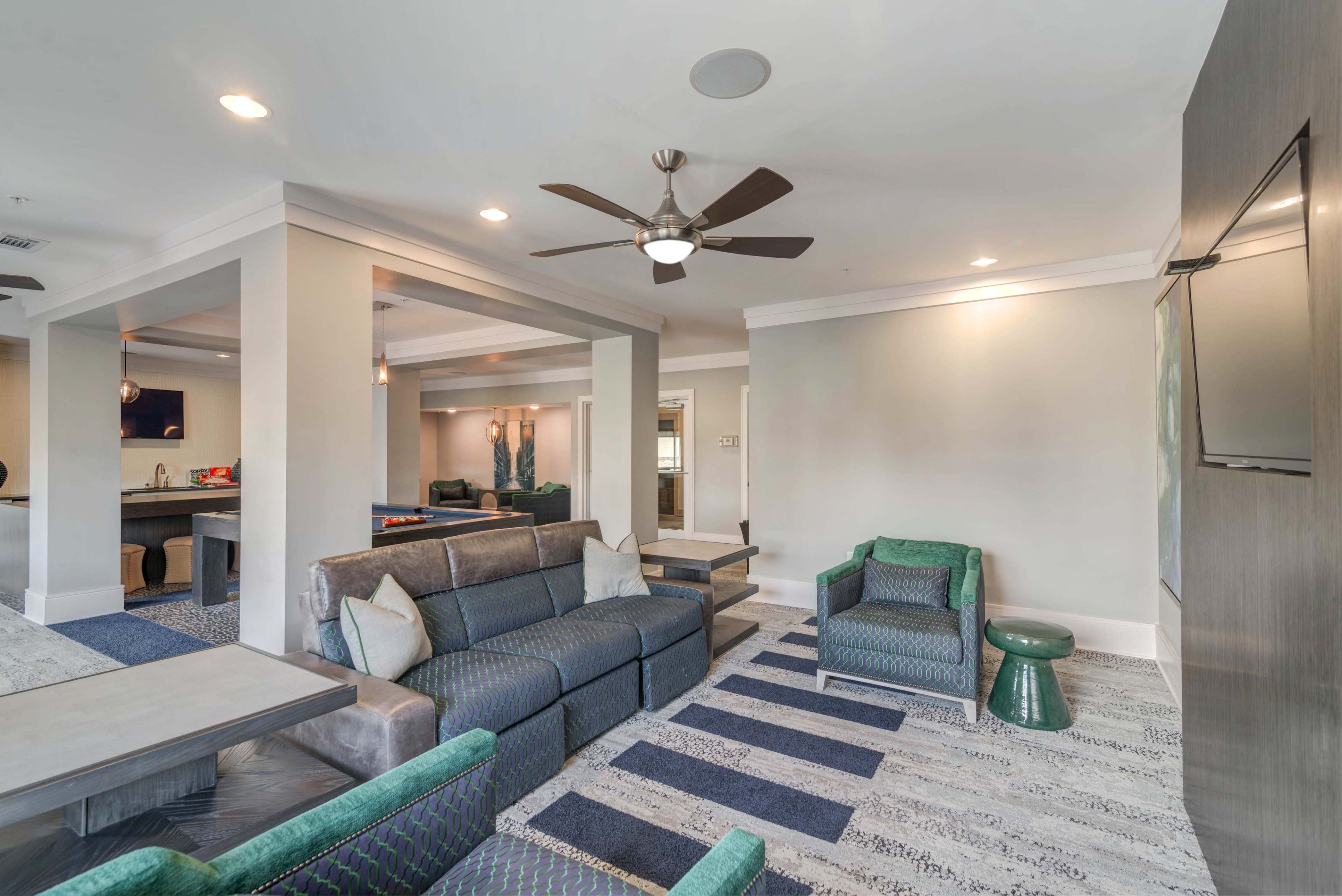 The image depicts a modern, open living area furnished with a gray sofa, patterned chairs, a ceiling fan, and a flat-screen TV on the wall.