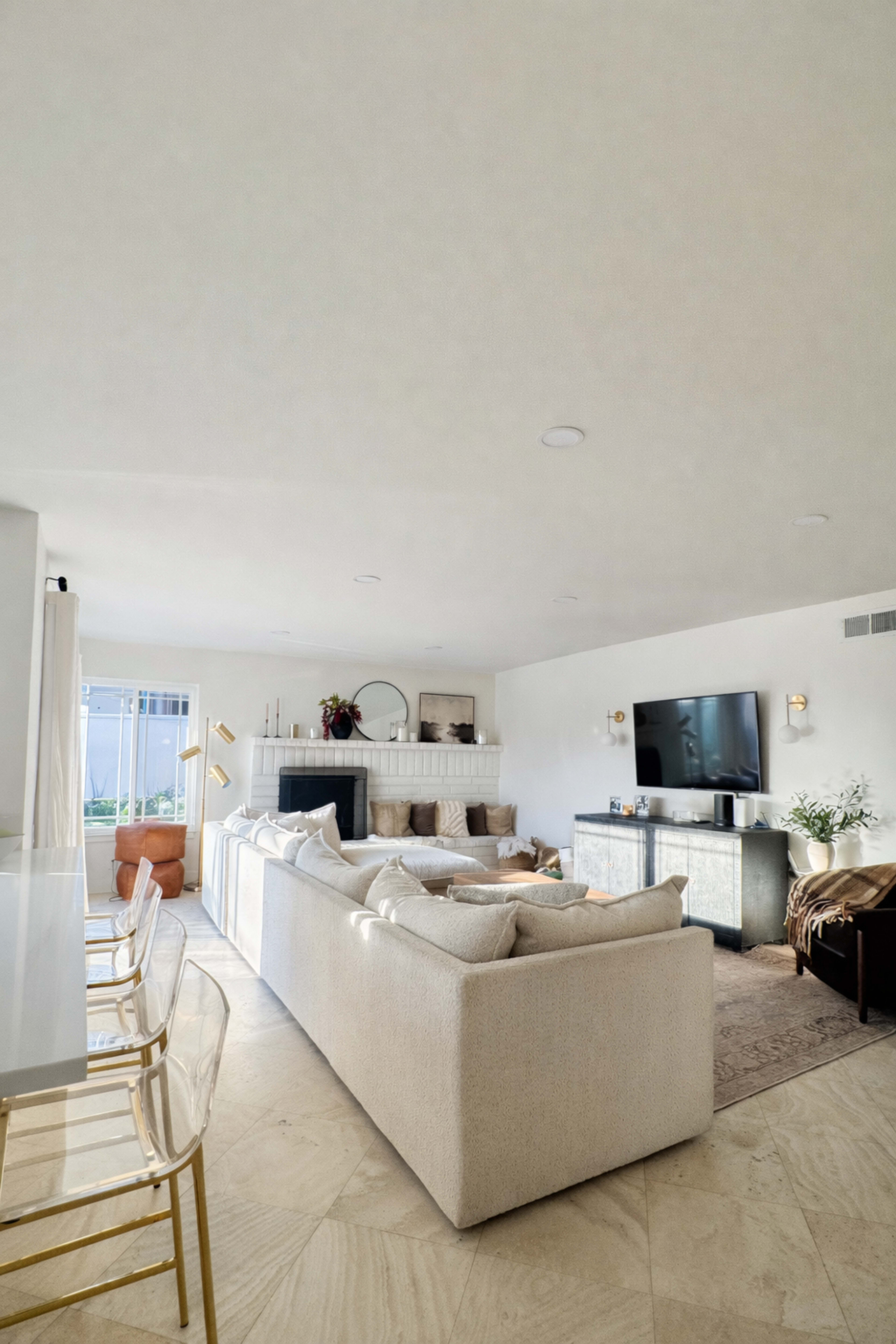 The image shows a modern living room with a sectional sofa, a TV mounted on the wall, and a fireplace in a light-filled space.