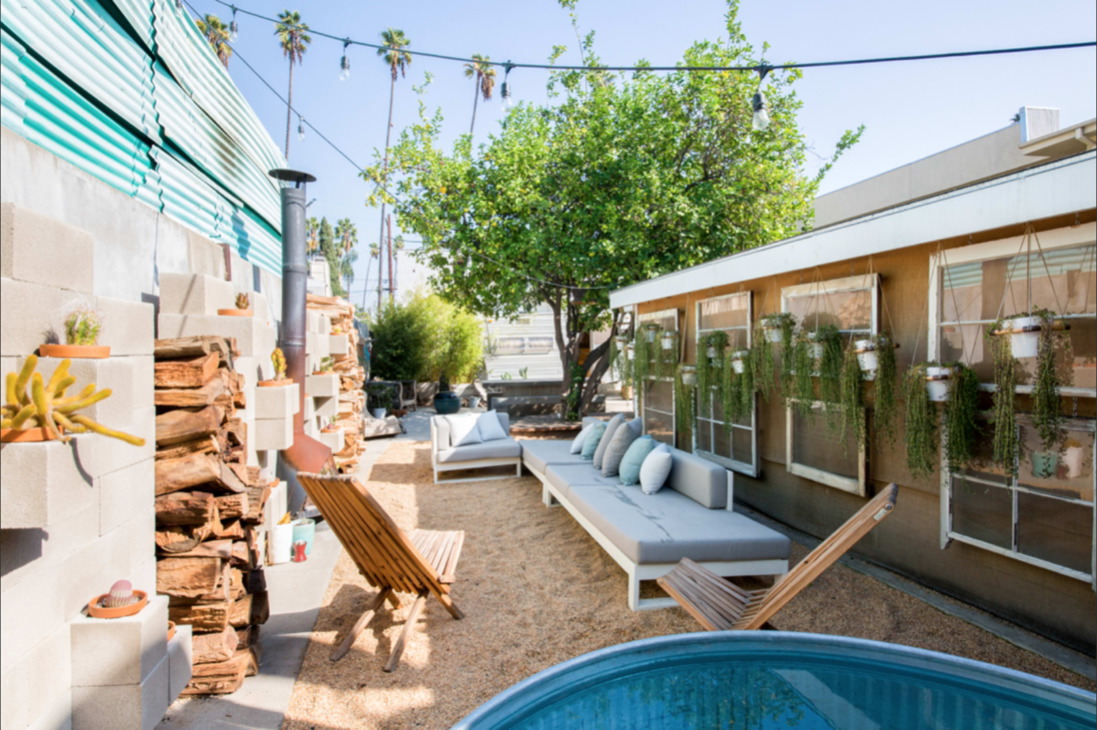 A small outdoor space with seating, a blue pool, green plants, stacked firewood, and a wall featuring decorative plant holders.
