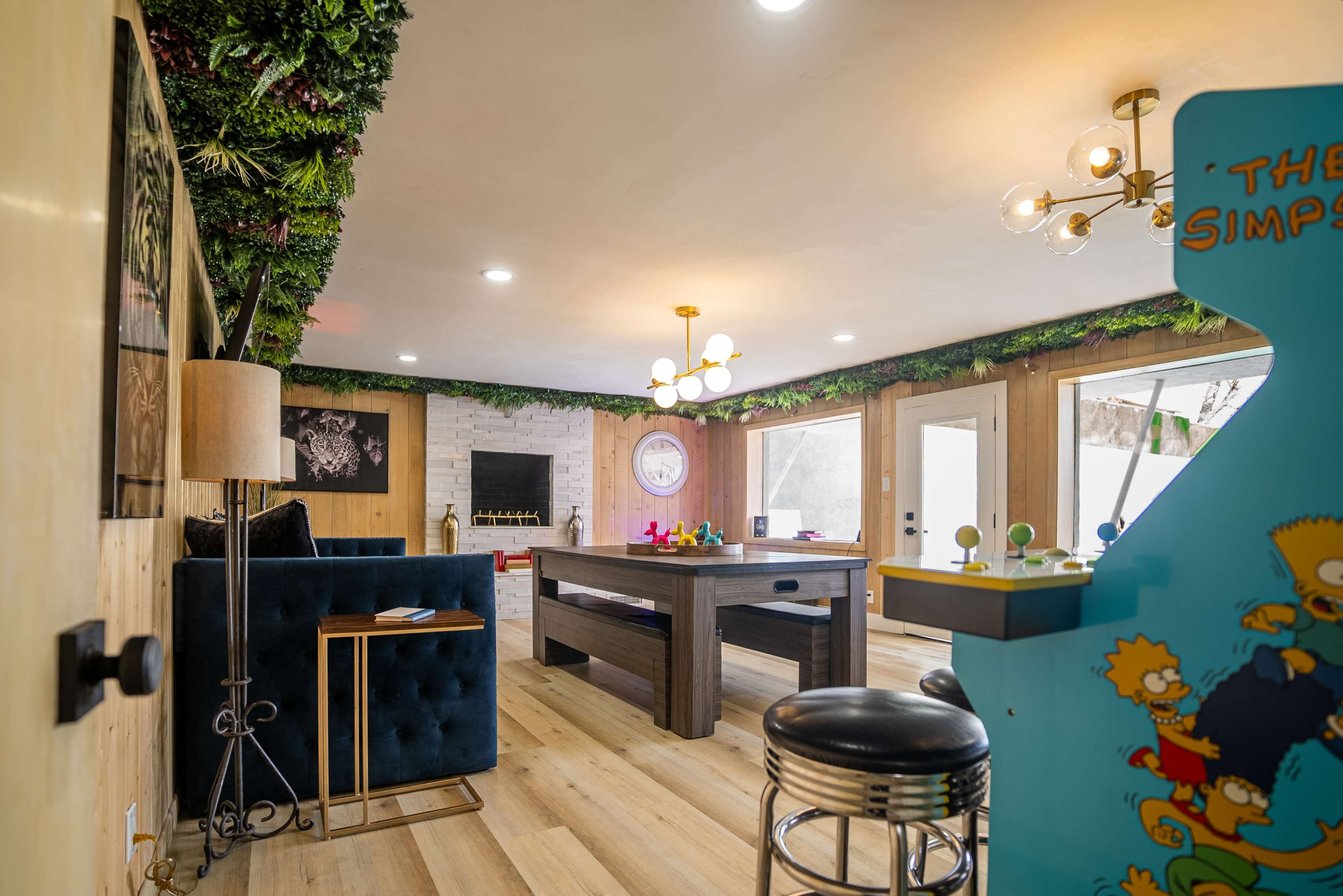 The image shows a modern interior room with a blue velvet couch, a large wooden table, and an arcade machine featuring characters from "The Simpsons."