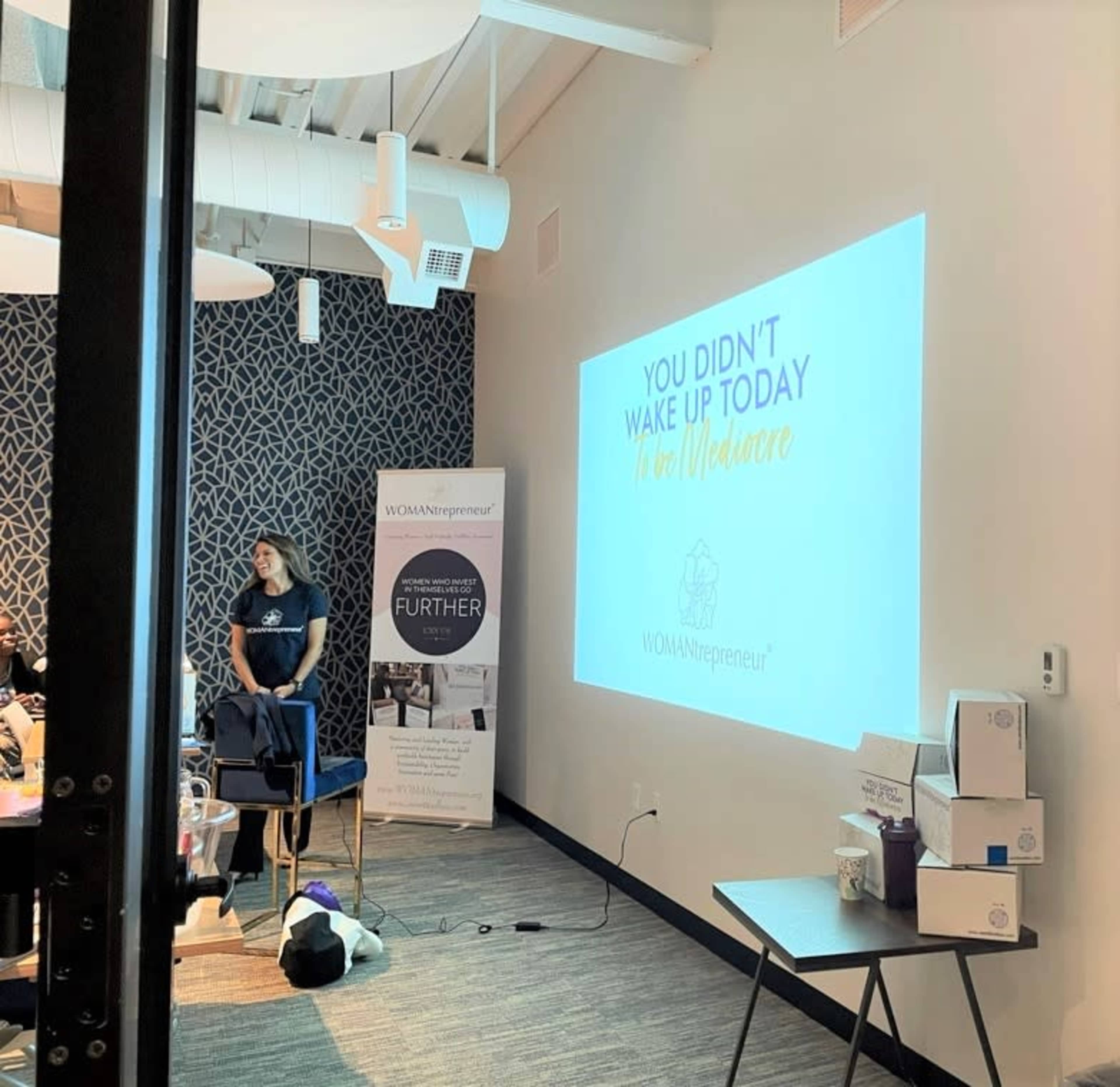 A woman stands beside a projector screen displaying the message "YOU DIDN'T WAKE UP TODAY TO BE MEDIOCRE" at a WOMANpreneur event.