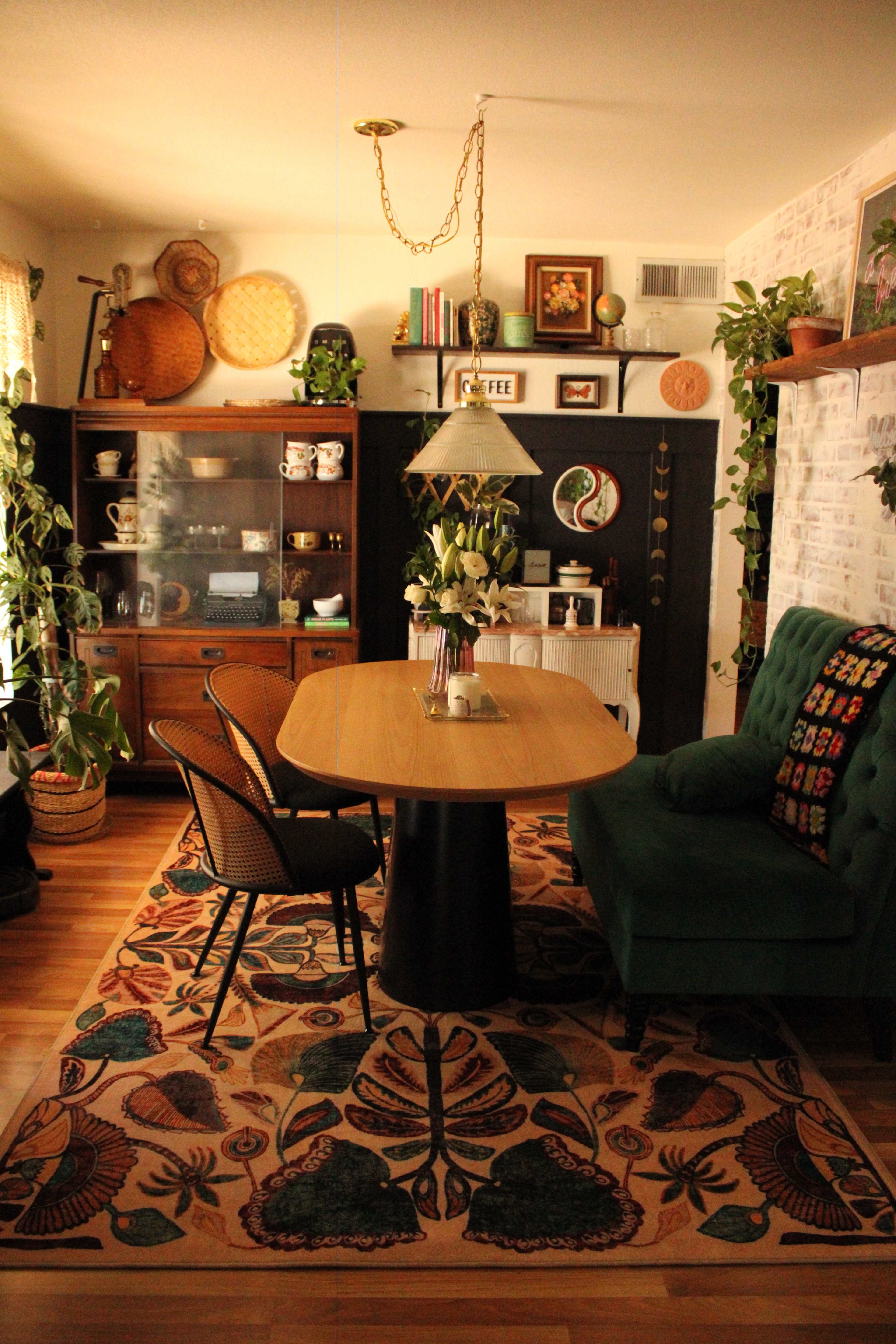 A cozy dining area featuring a round wooden table surrounded by chairs, a green upholstered sofa, and various plants and decorations on the walls and shelves.