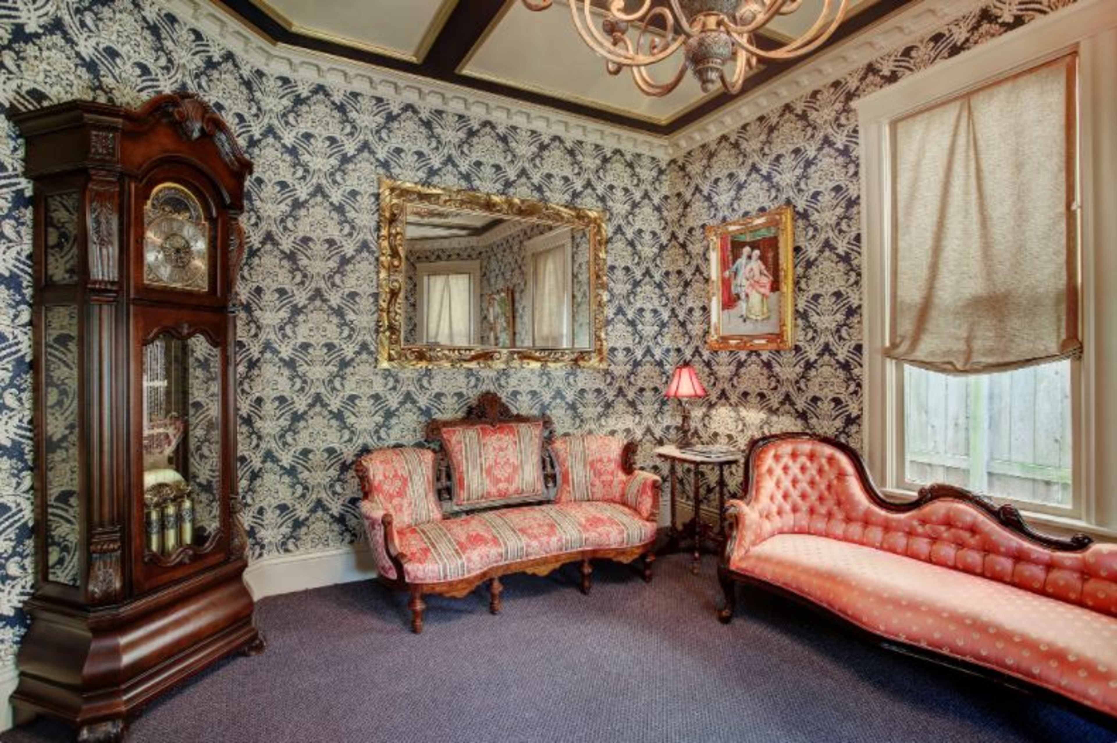 A vintage parlor featuring a floral wallpaper, a large mirror, a grandfather clock, and two upholstered sofas.