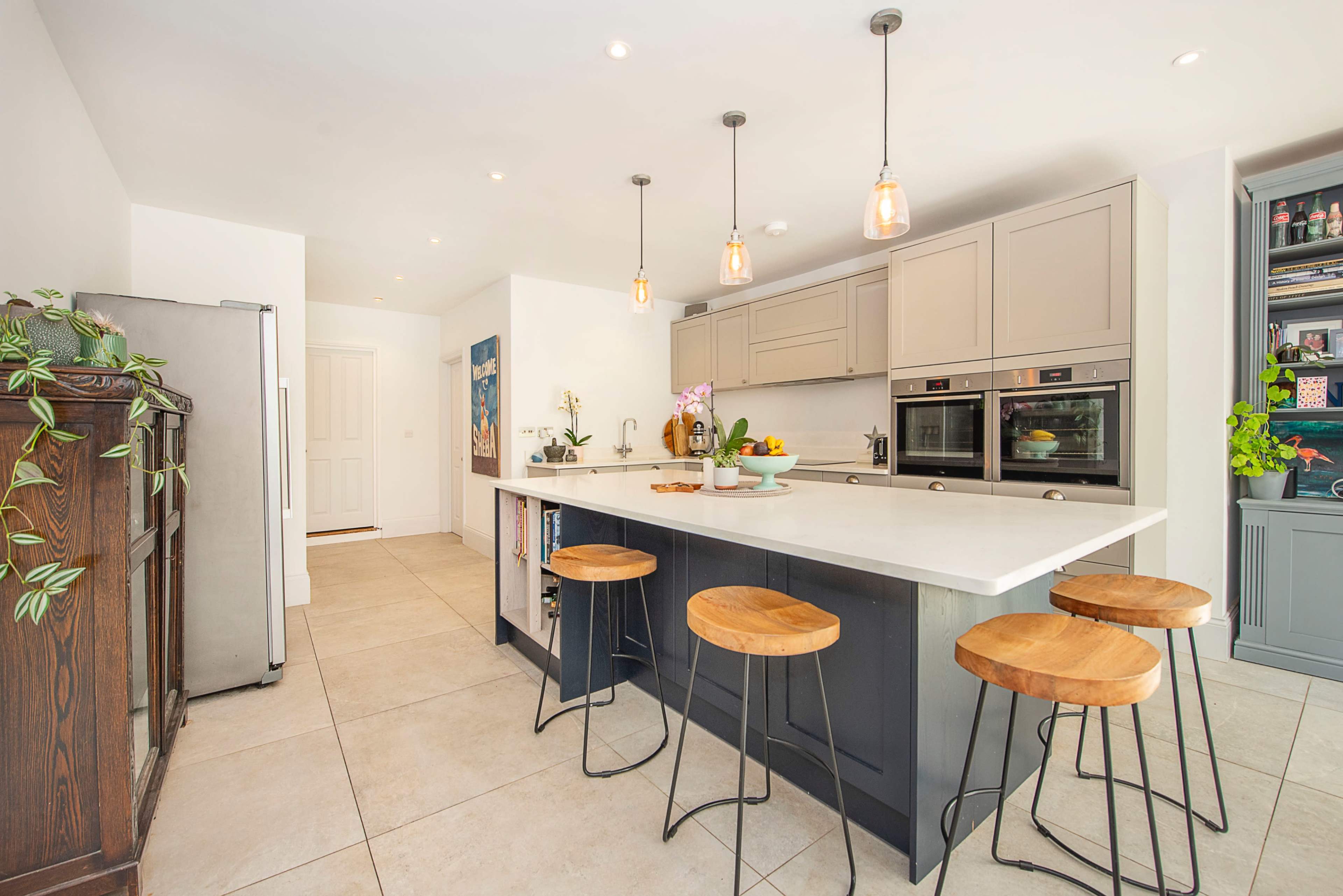 The image shows a modern kitchen featuring a central island with four wooden stools, built-in appliances, and light fixtures hanging from the ceiling.