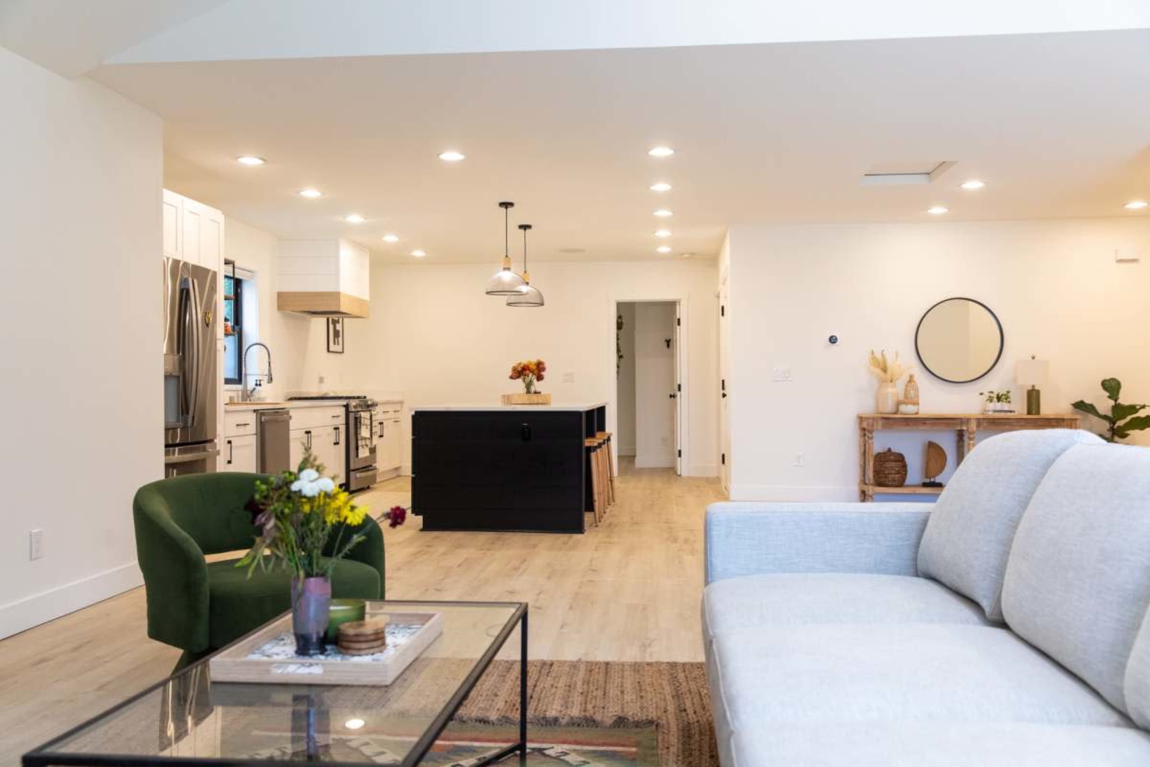 The image shows a modern living space featuring a gray sofa, a green chair, and an open kitchen with white cabinetry and a central island.