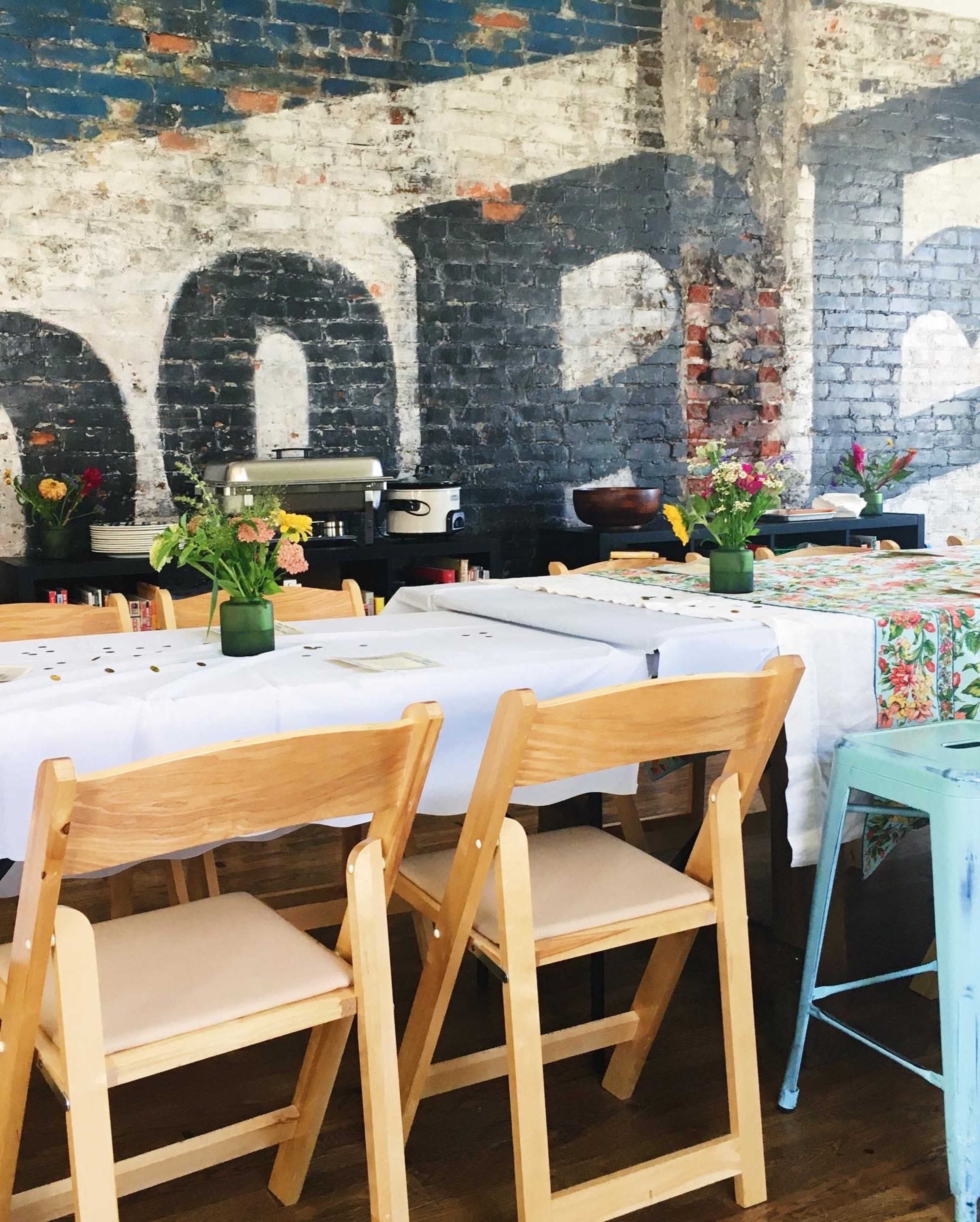 A rustic dining area with wooden folding chairs arranged around tables covered in white and patterned cloths, and small flower vases on each table.