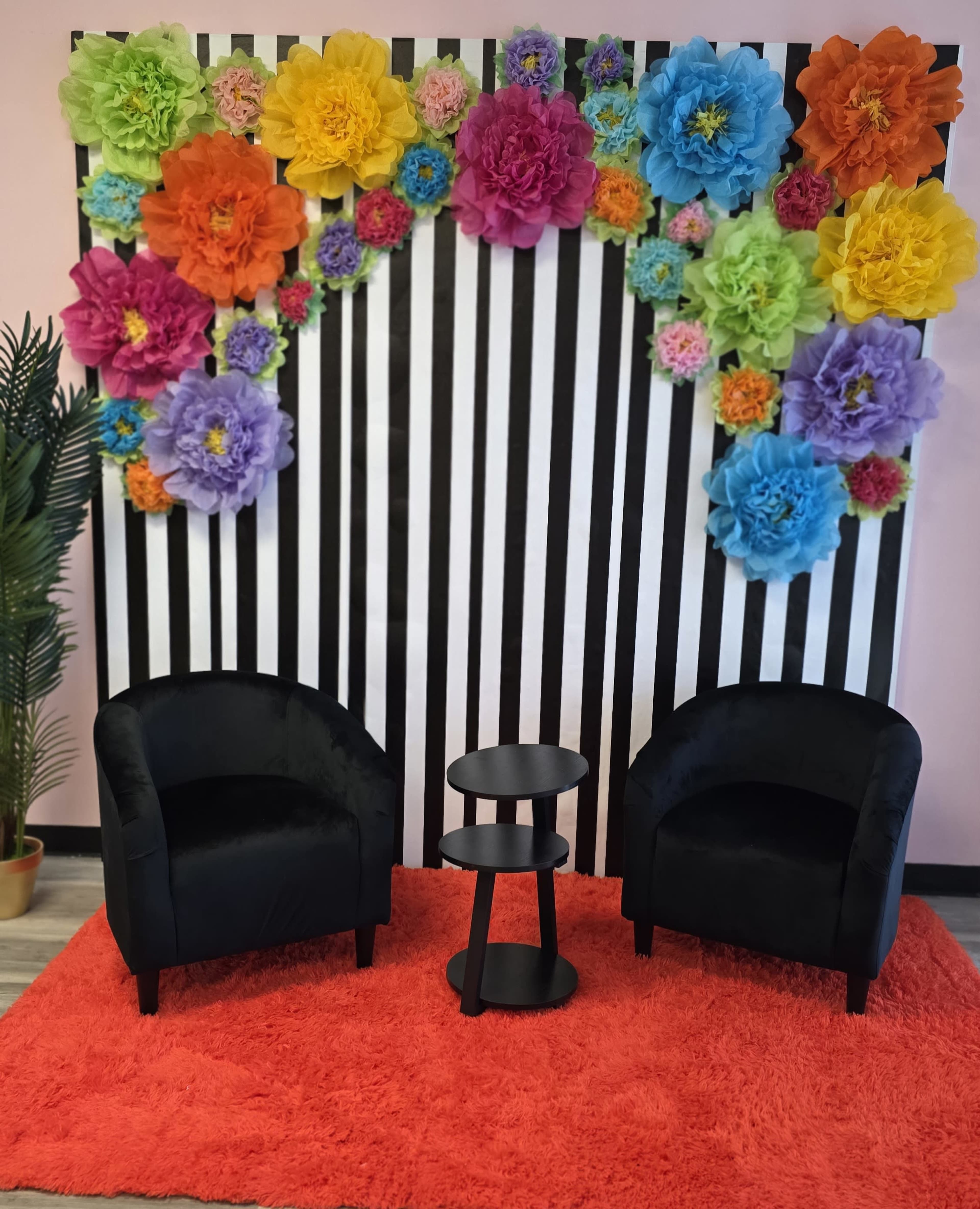 The image shows a brightly decorated backdrop of large paper flowers in various colors against a black and white striped wall, with two black chairs and a small black table on a red fuzzy rug.