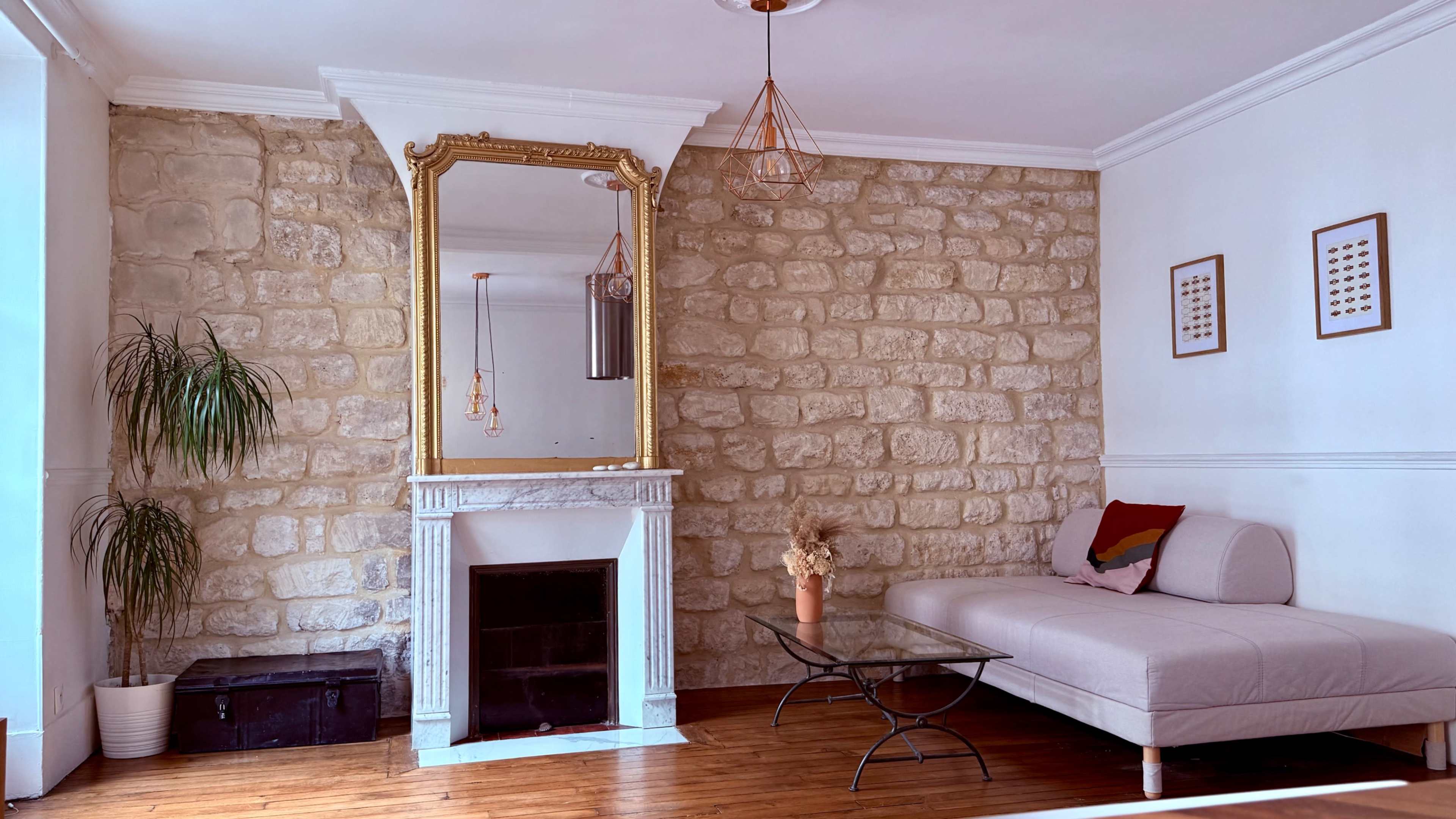 The image shows a living room with a stone wall, a large mirror in a gold frame, a glass coffee table, a light-colored sofa, and decorative elements like a plant and wall art.