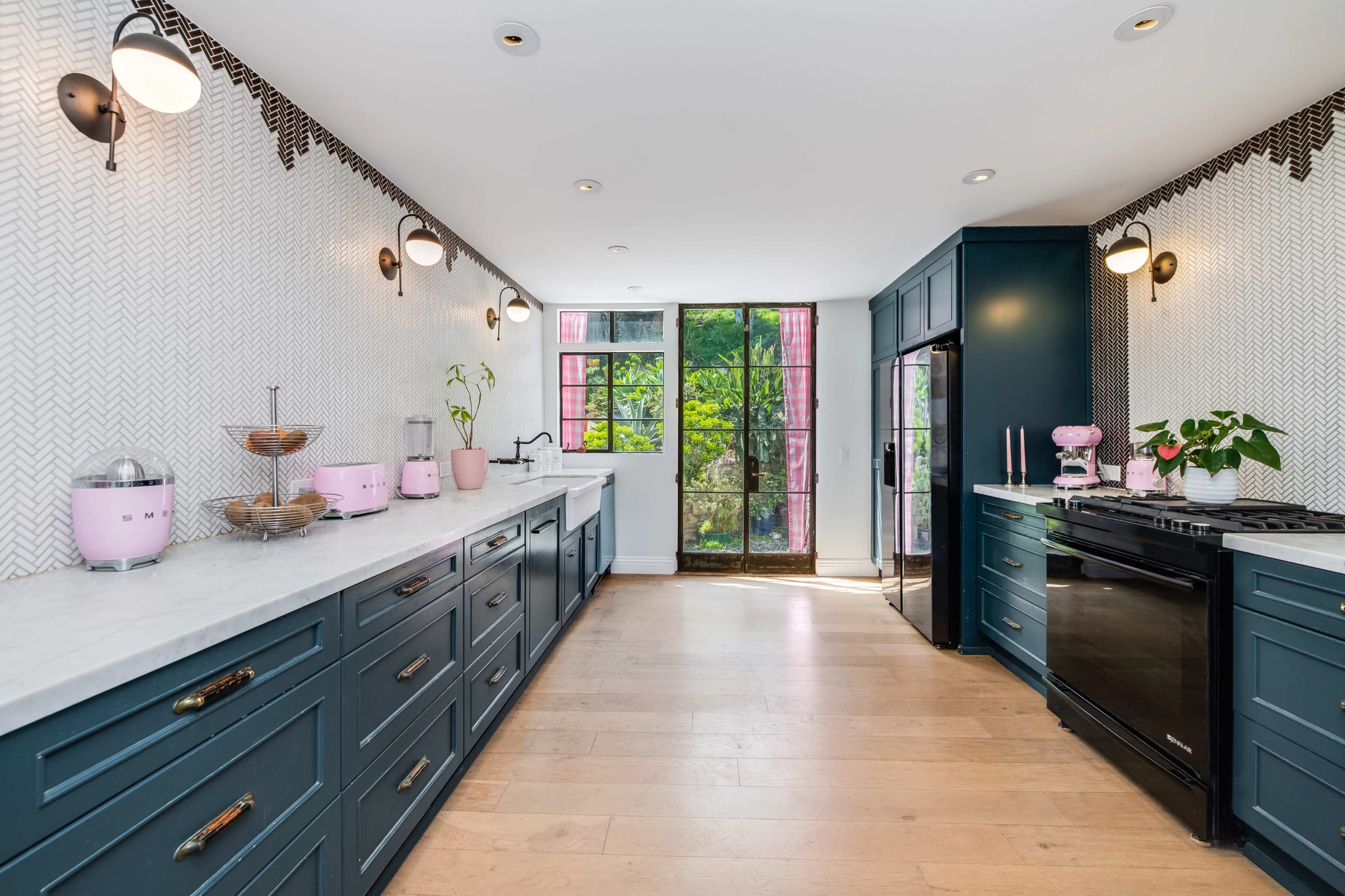 The image shows a modern kitchen featuring dark blue cabinetry, white countertops, and large windows overlooking greenery.