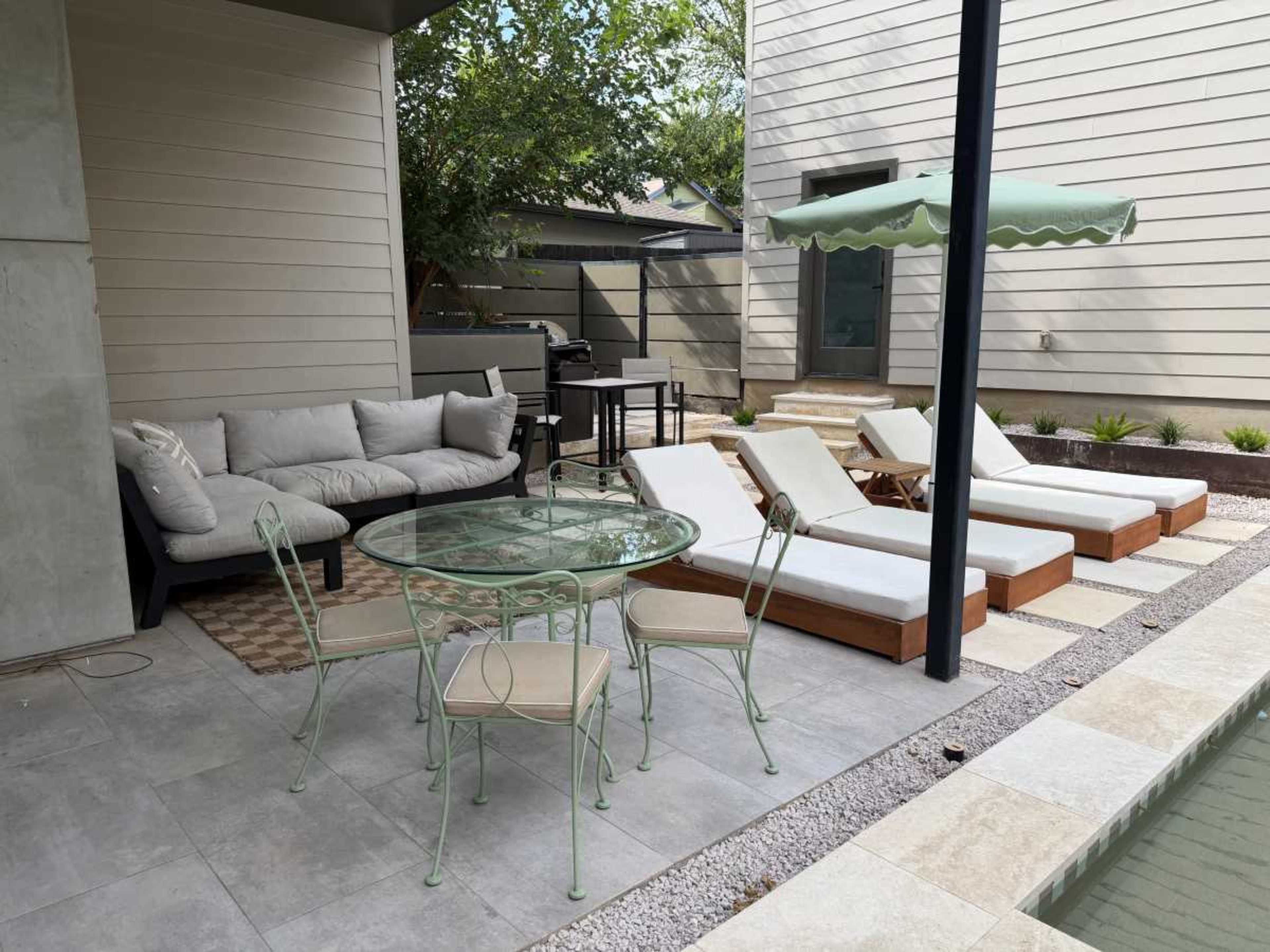 The image shows a patio area with a glass table surrounded by green metal chairs, a sectional sofa, and two lounge chairs beside a pool, alongside a grey wall and a green umbrella.