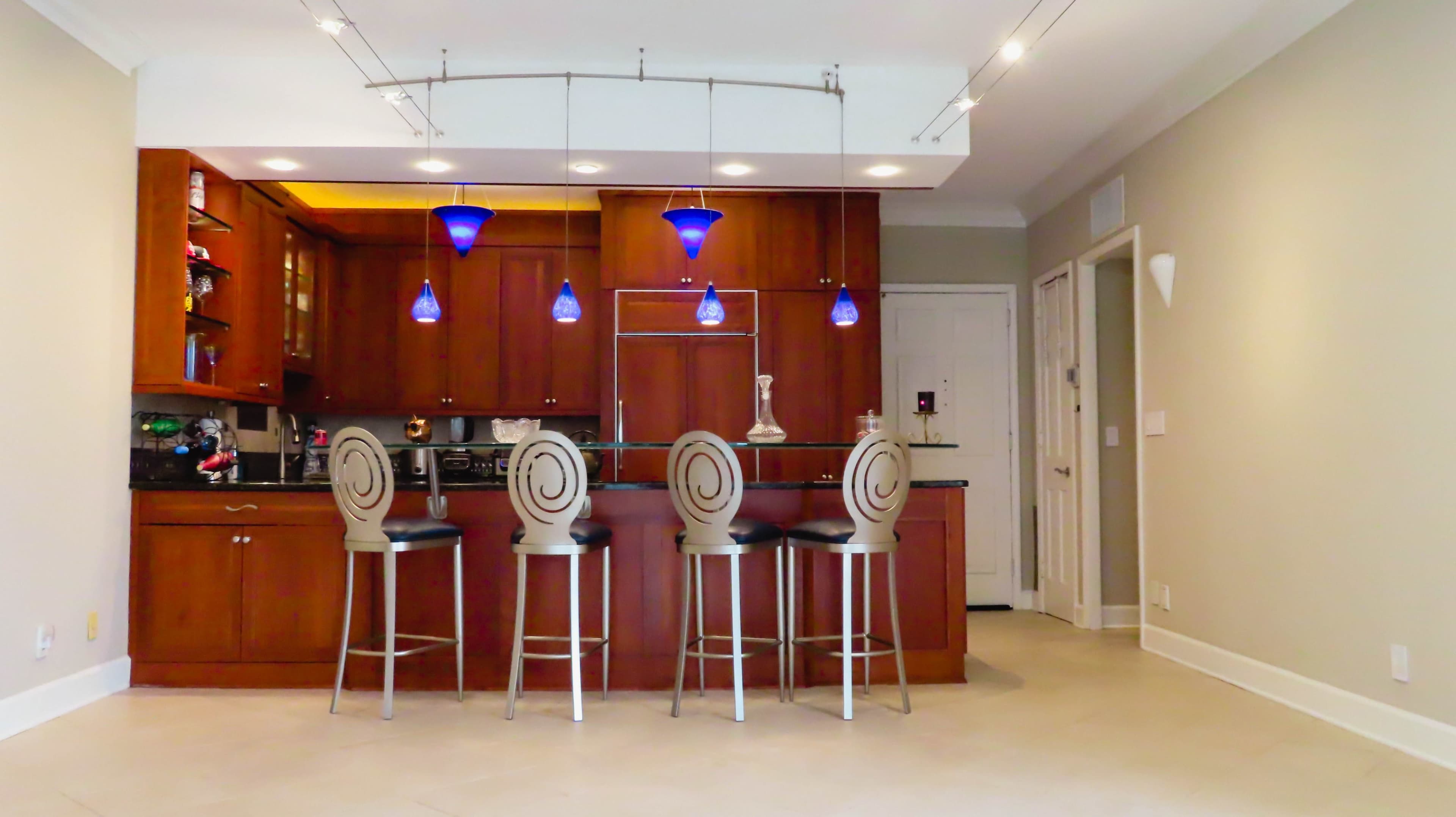 A modern kitchen with wooden cabinetry, a bar counter with four stools, and blue pendant lights above.