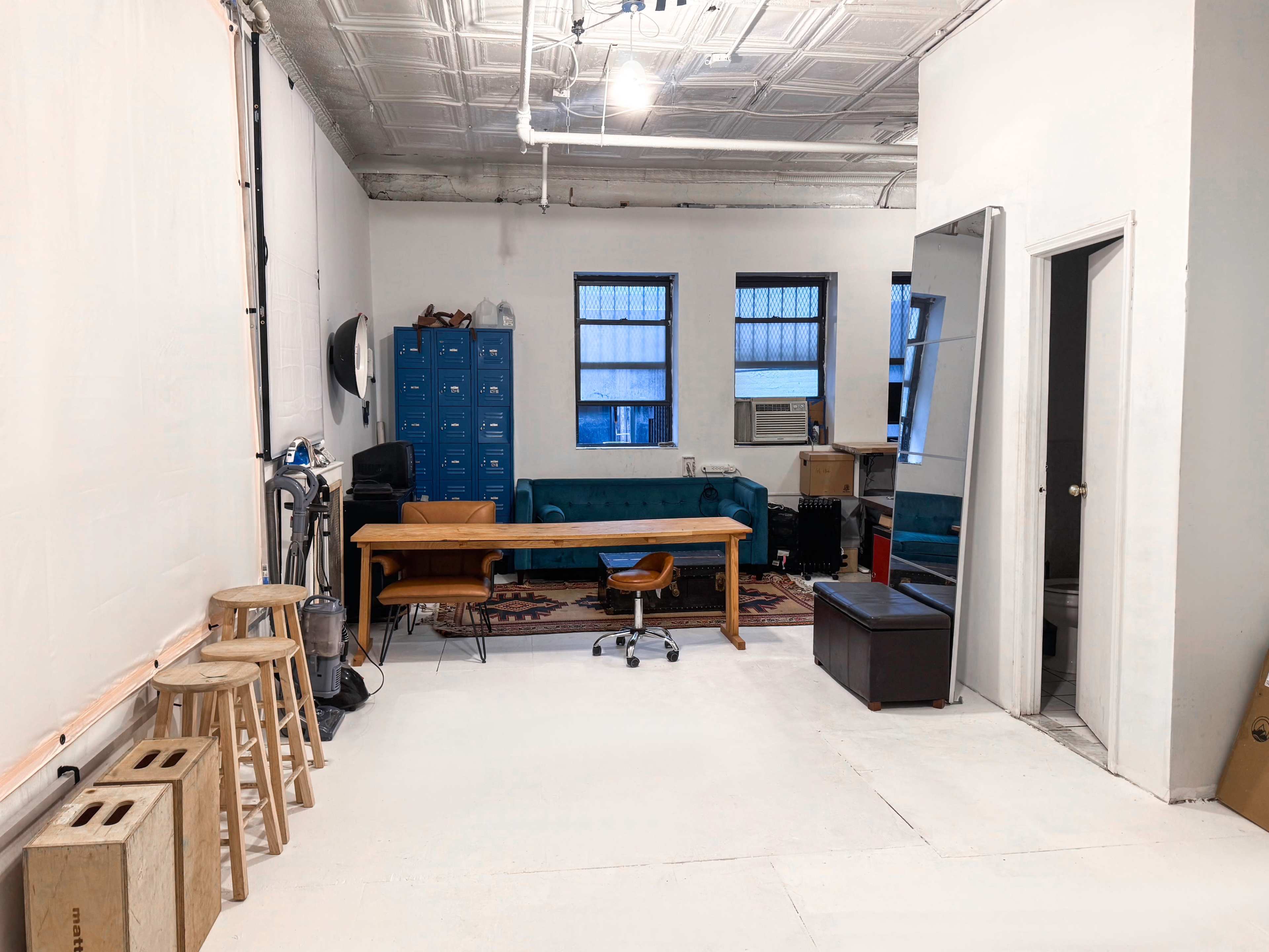 The image shows a spacious room with light-colored walls, a green couch, a wooden table with a chair, and storage lockers against one wall.