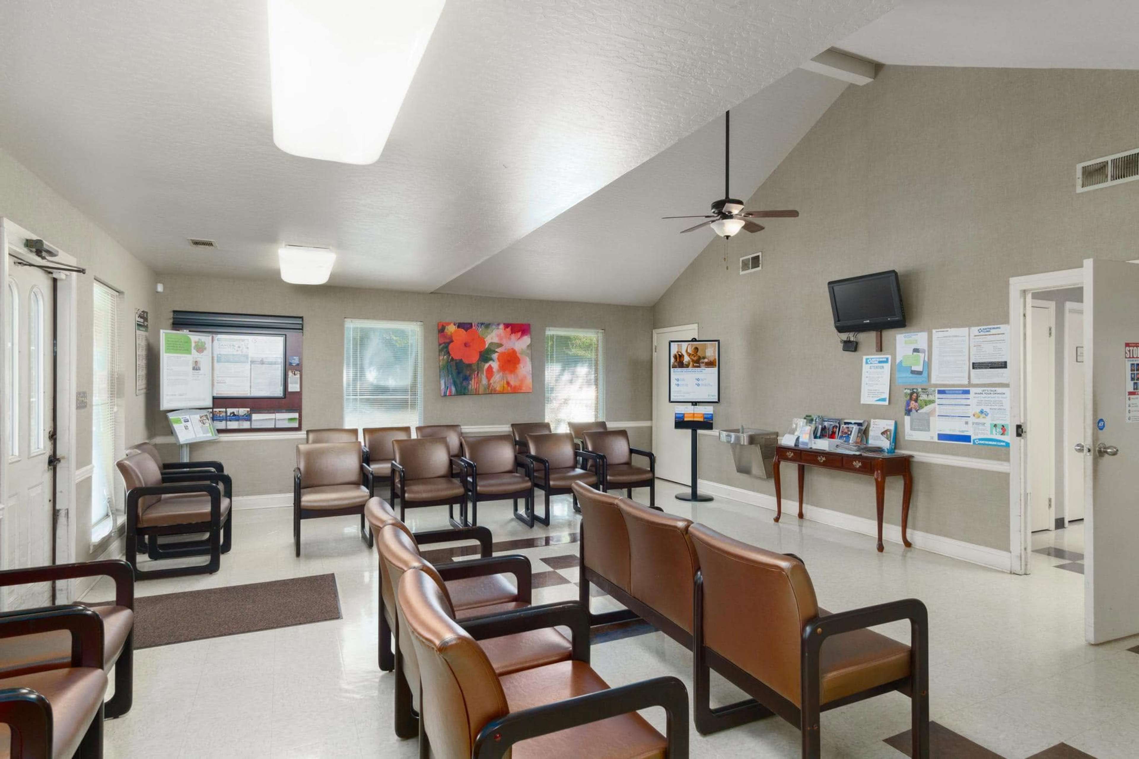 The image shows a waiting room with several brown chairs arranged in rows, a television mounted on the wall, and informational posters displayed.