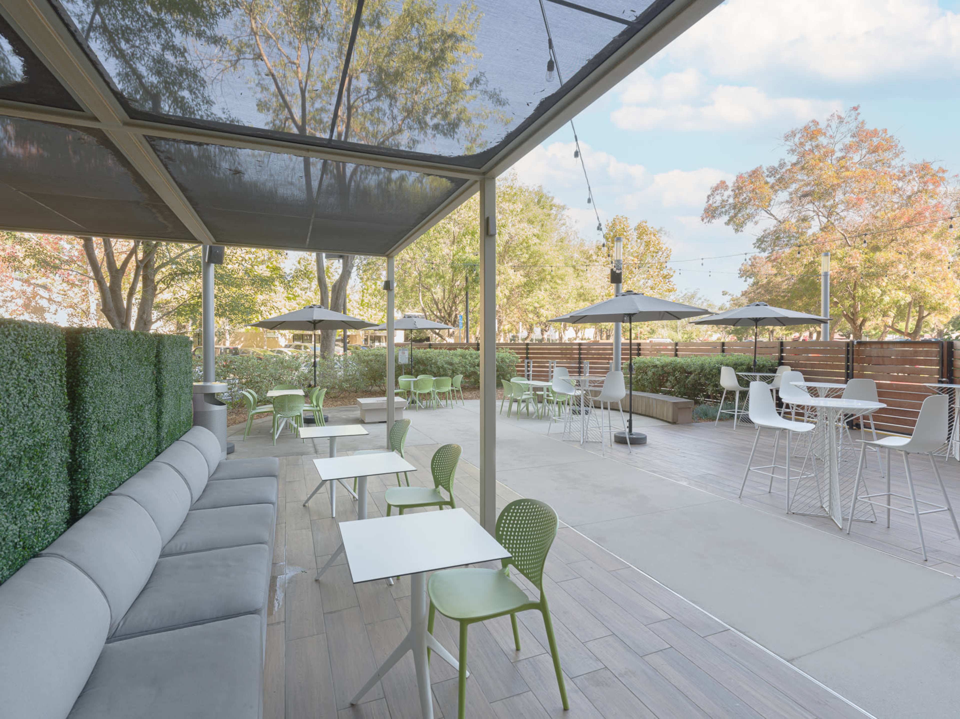 The image shows a modern outdoor café area with green furniture, tables, and umbrellas surrounded by trees.