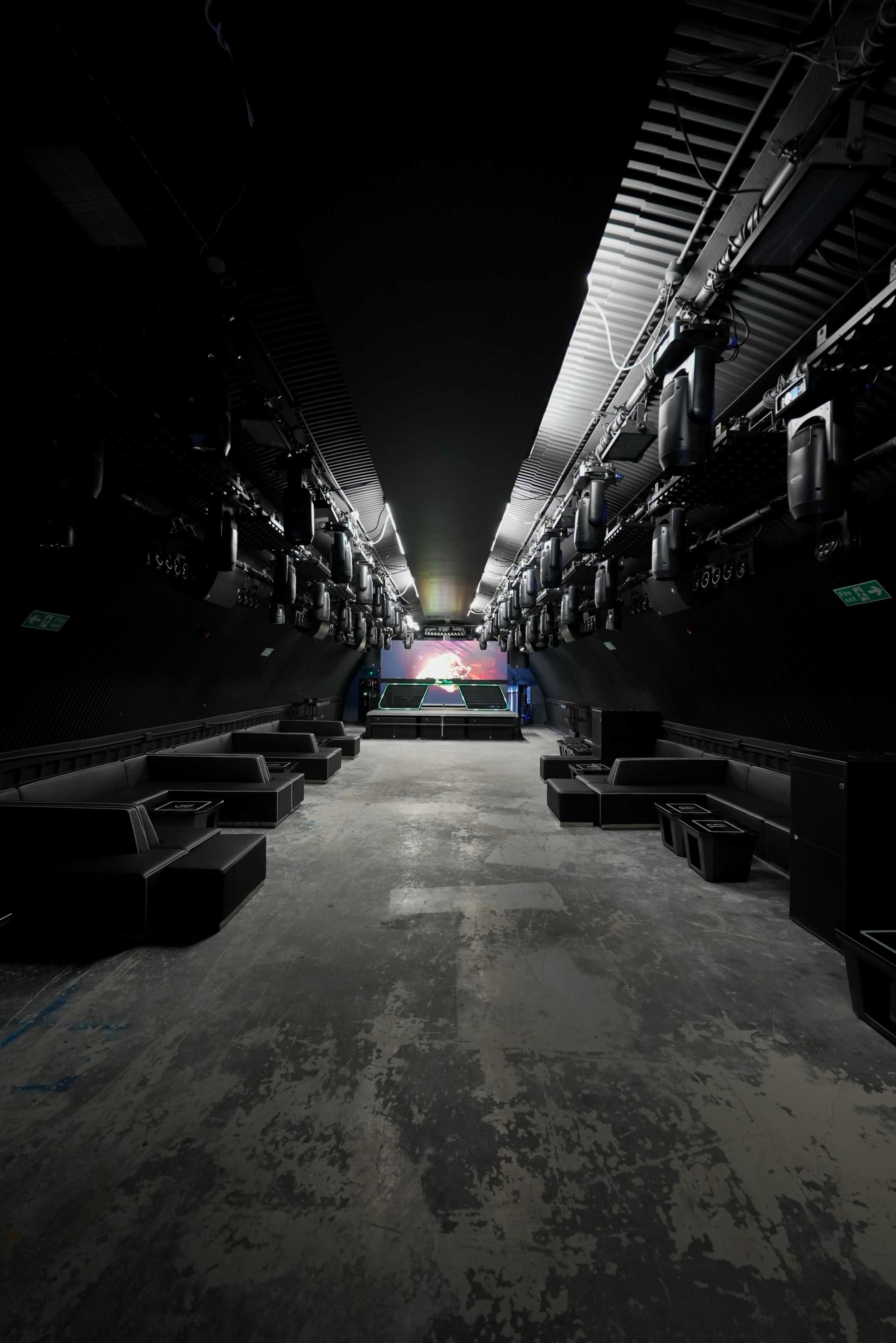 The image shows a long, dimly lit indoor space with a stage at the far end and several rows of black seating along the sides.