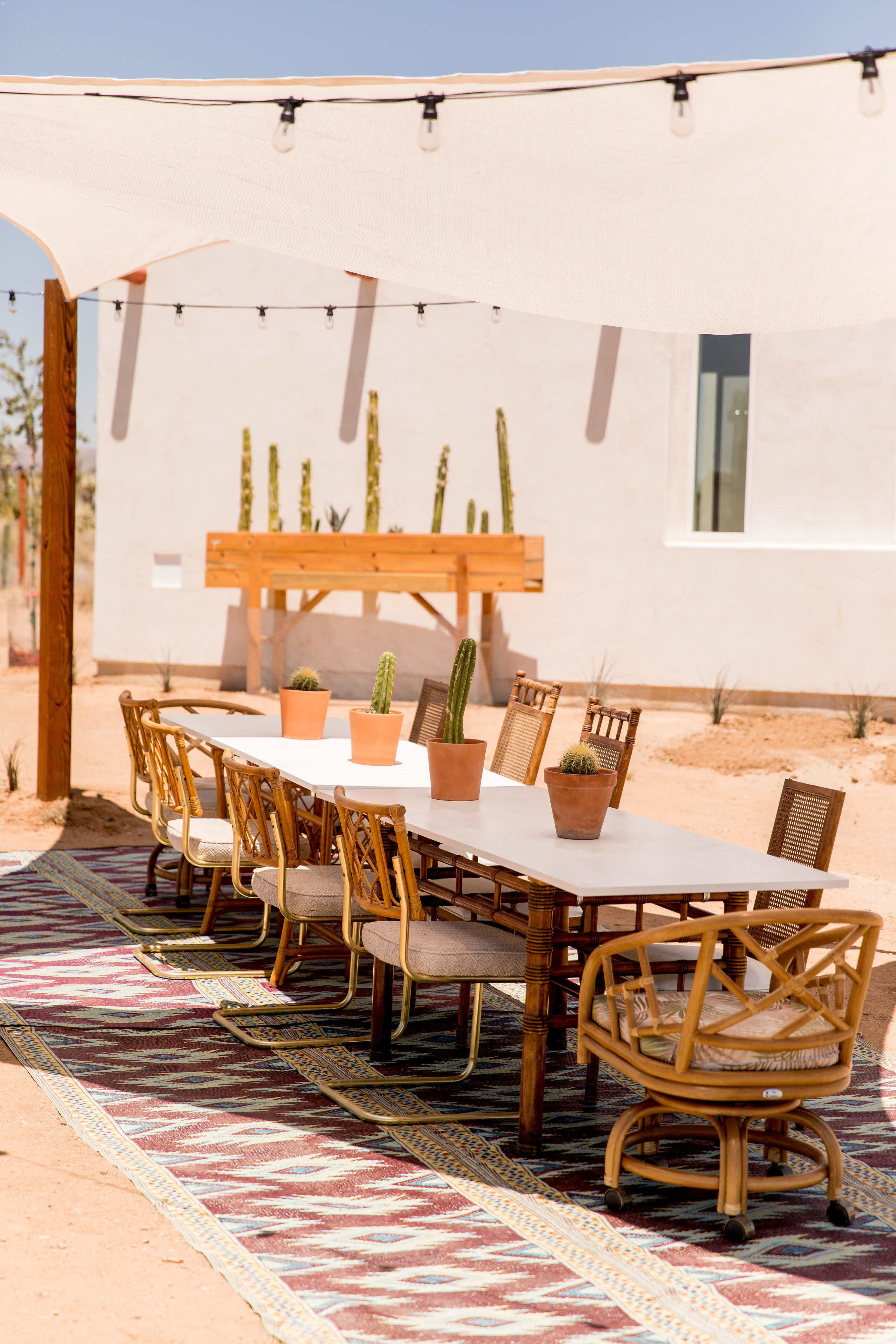 A long, white table with chairs is set up outdoors under a shaded area, surrounded by potted cacti and a patterned rug.