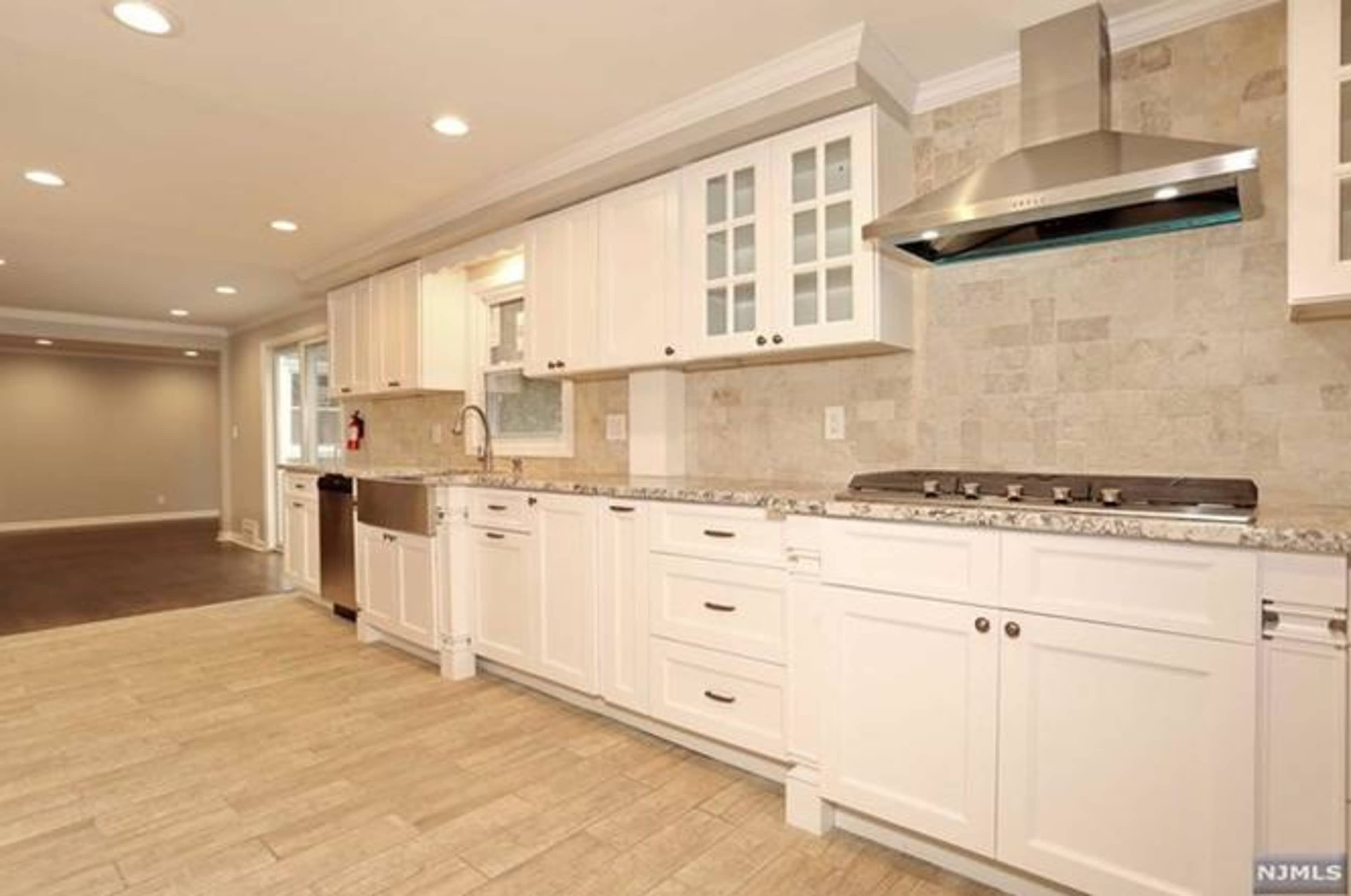 A modern kitchen with white cabinets, a stainless steel range hood, and a tiled backsplash, adjacent to a spacious living area.
