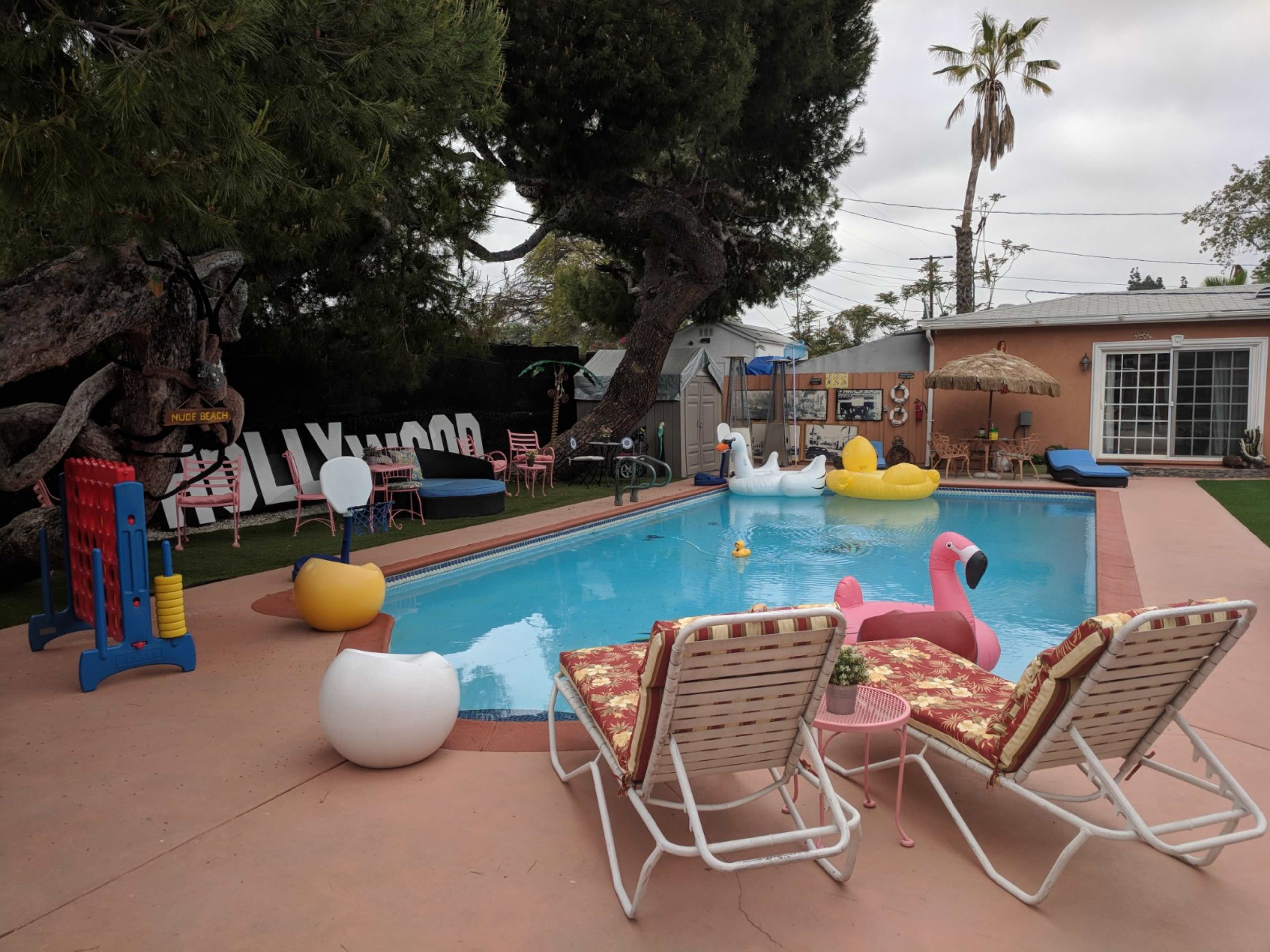 The scene features a backyard with a swimming pool surrounded by colorful inflatable pool floats, two lounge chairs, and a shaded area with an umbrella.