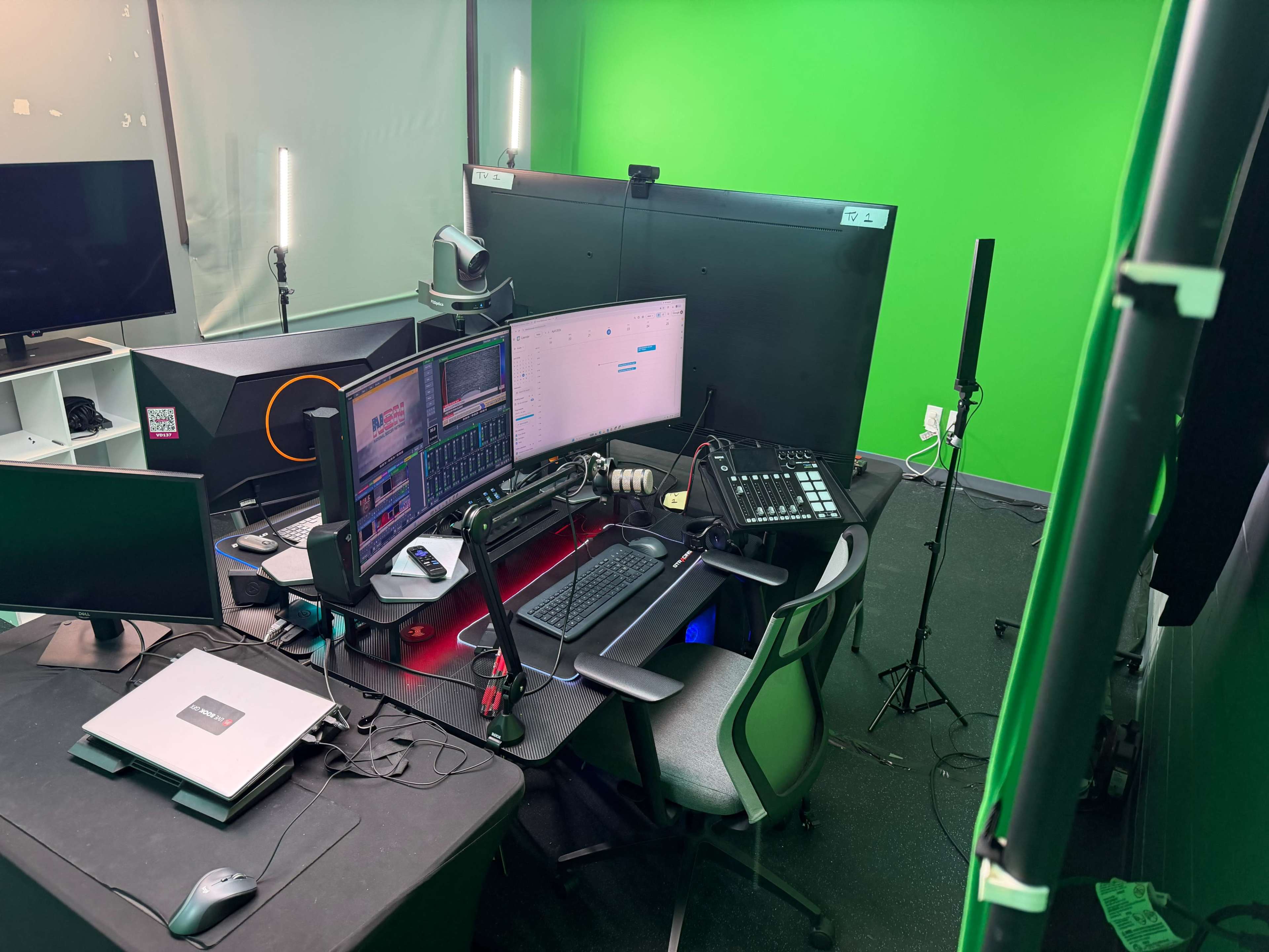 The image shows a workspace with multiple computer monitors, a mixing console, and a green screen in the background, set up for video production or streaming.