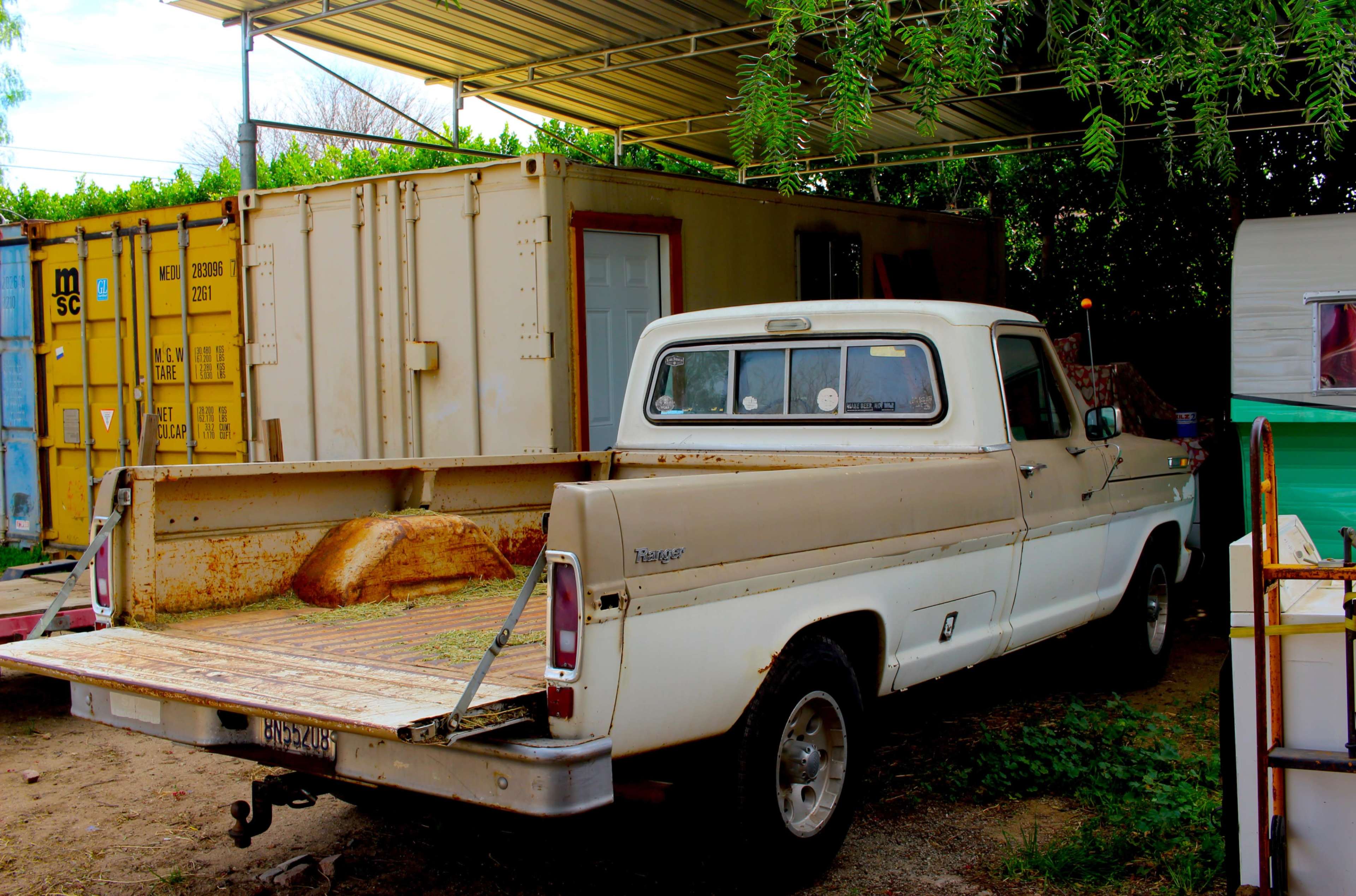 An old pickup truck is parked beside a shipping container and a trailer under a canopy.
