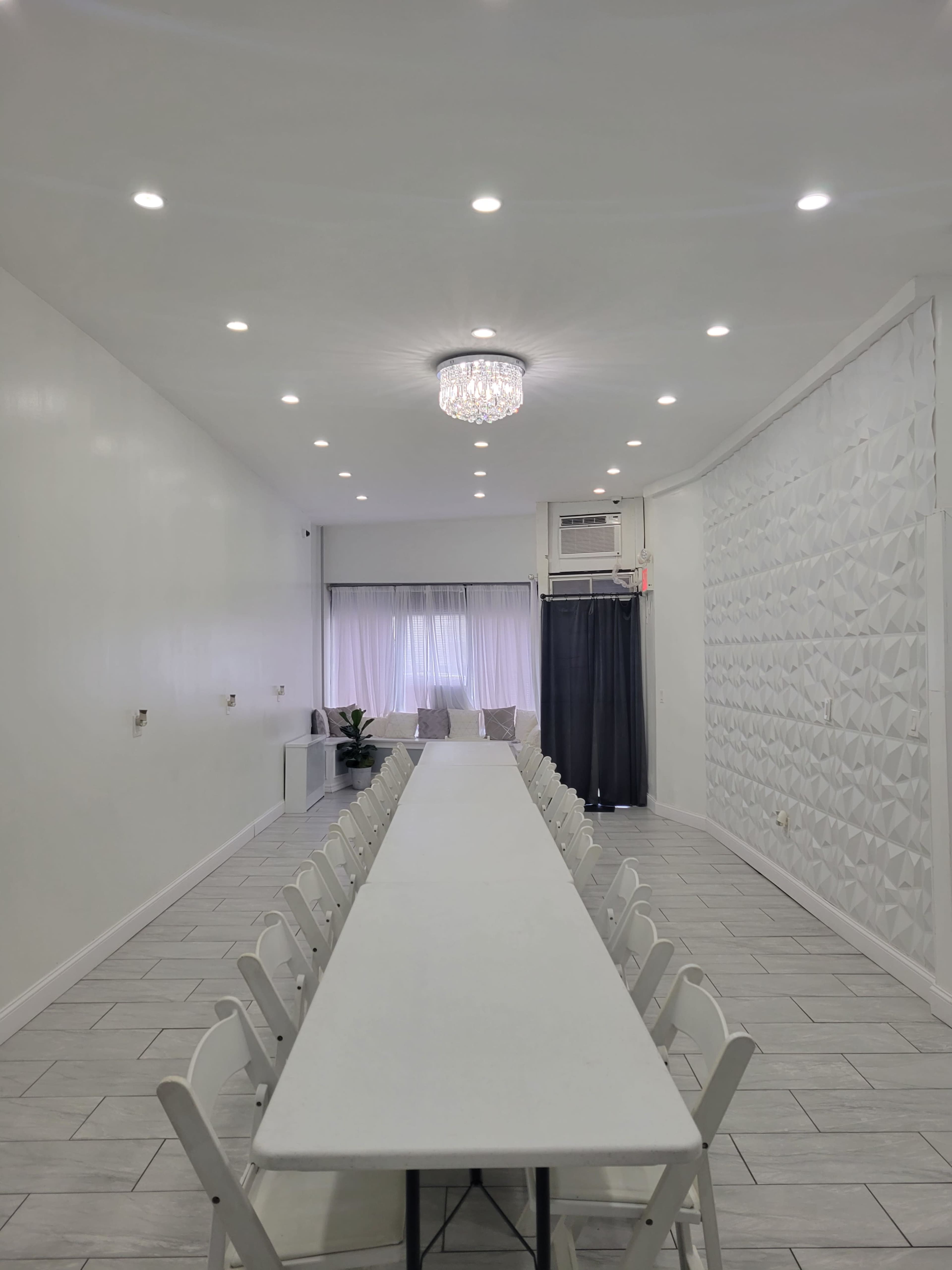 A long, white table is set in a well-lit room with a chandelier, featuring a textured wall and large windows at one end.
