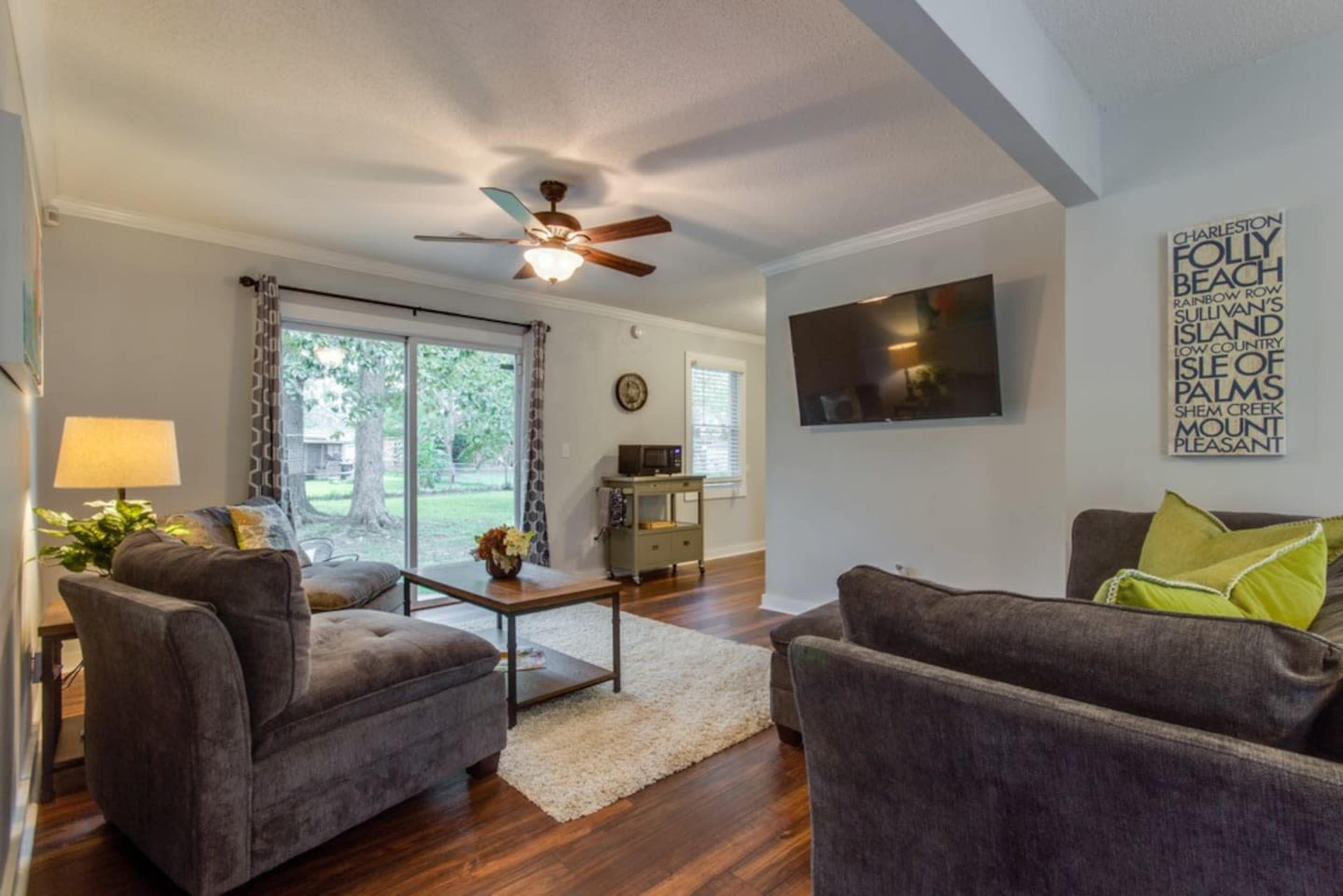The living room features a ceiling fan, a television mounted on the wall, a coffee table, and two gray sofas arranged around a rug.