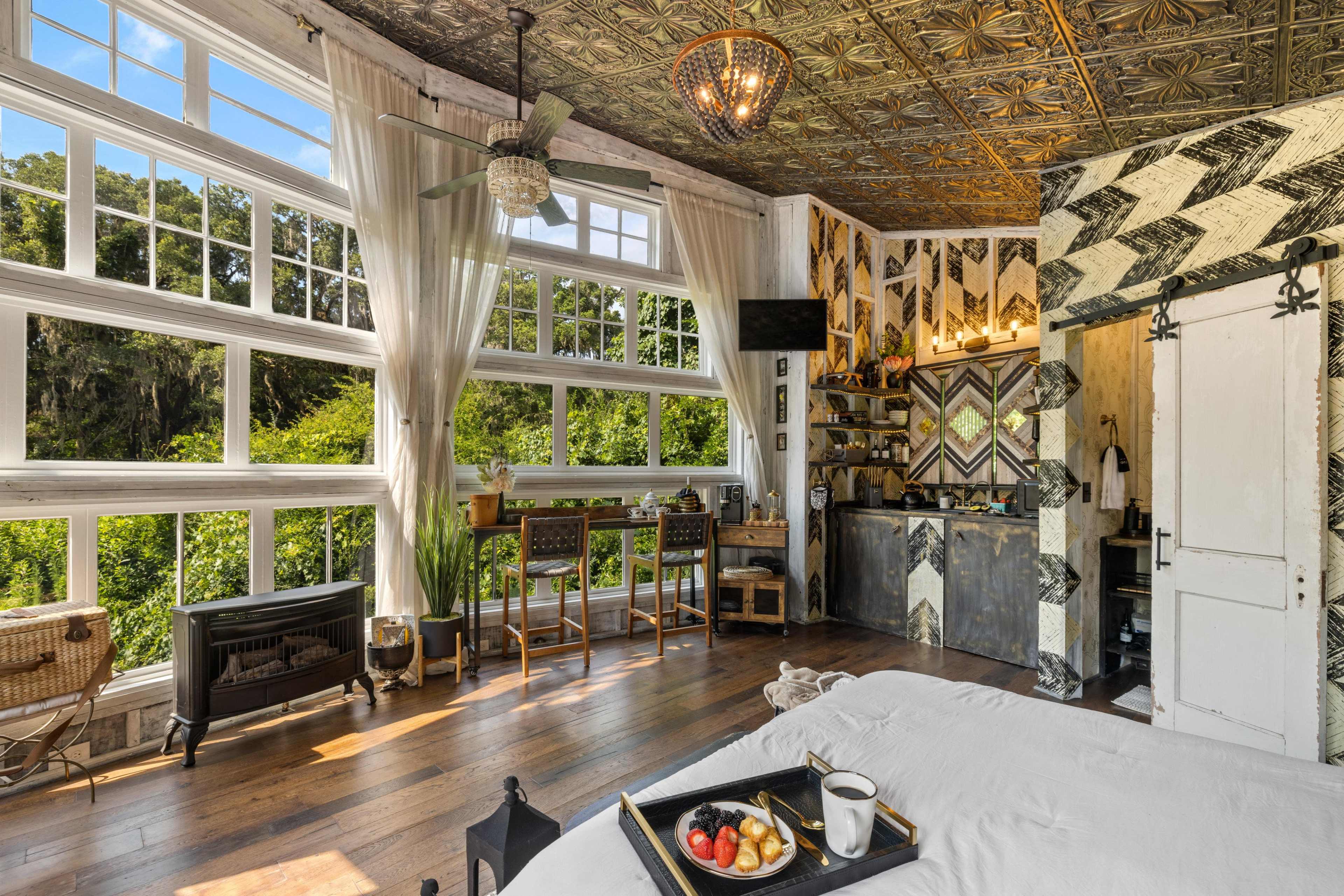 A bright bedroom features large windows, a kitchenette, and a seating area with wooden bar stools.