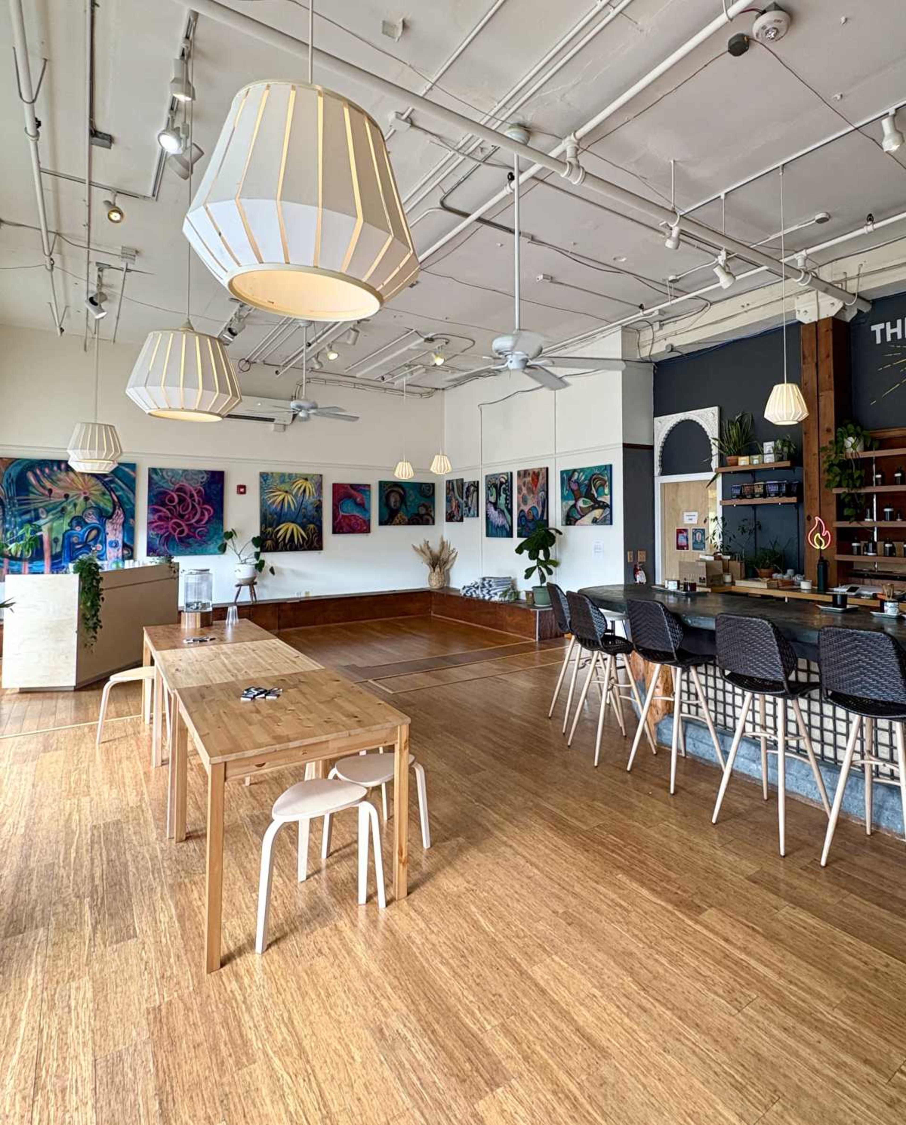The image shows a spacious café interior with modern wooden furniture, large pendant lights, and colorful artwork on the walls.