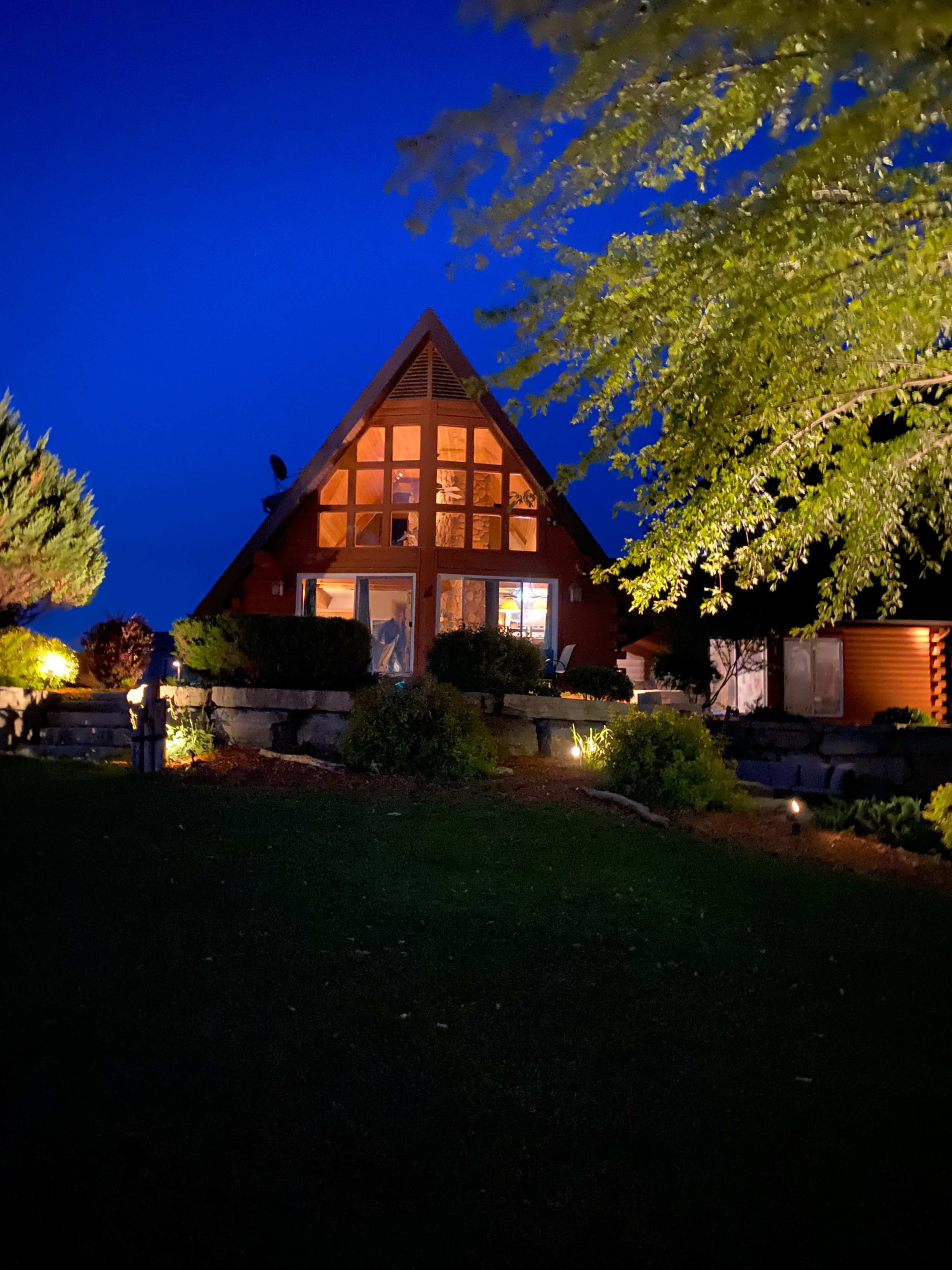 A triangular-roofed house with large front windows is illuminated at night, surrounded by landscaped gardens and trees.