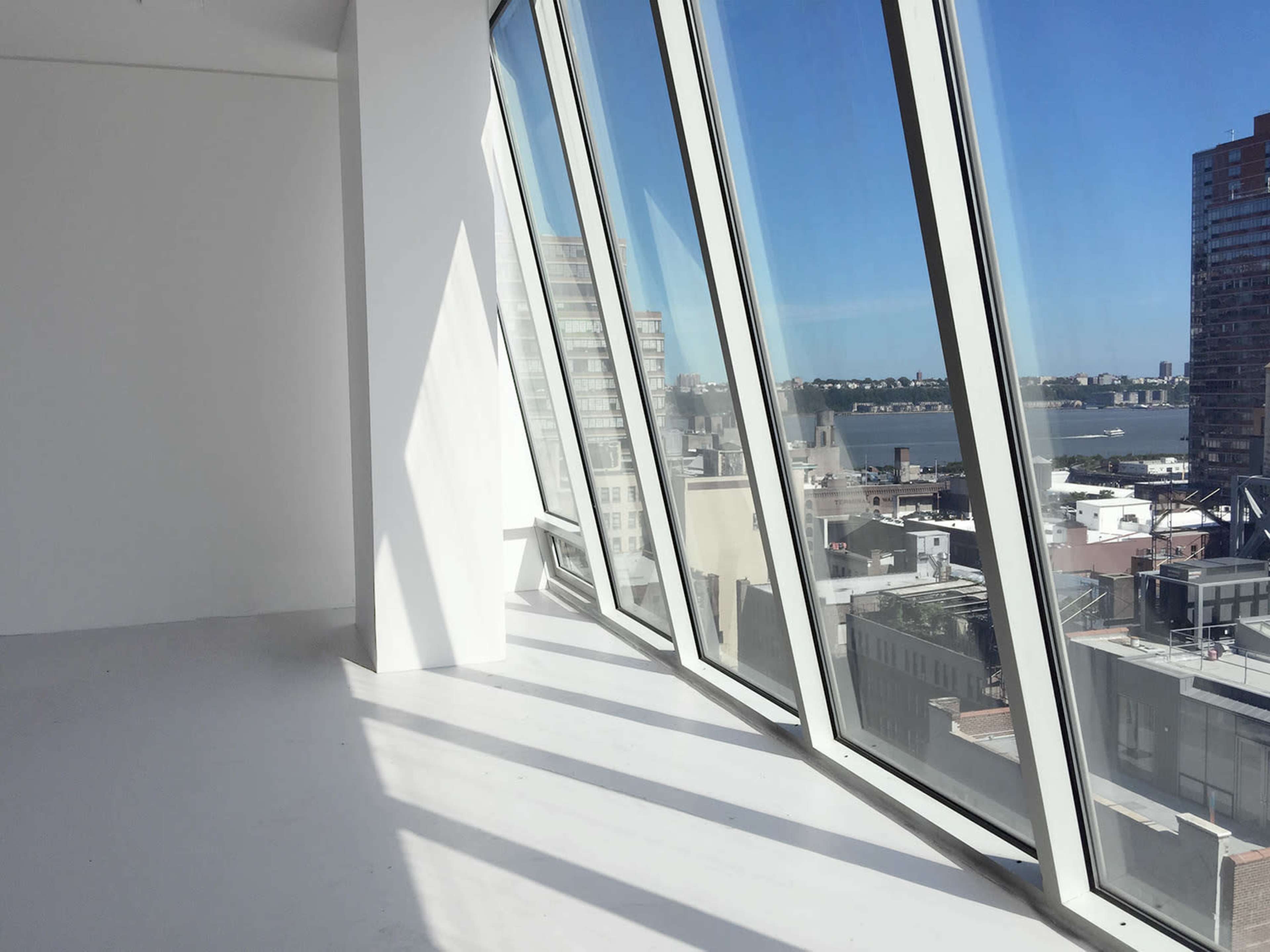 A spacious room with large, angled windows offers a view of the city skyline and river beyond.