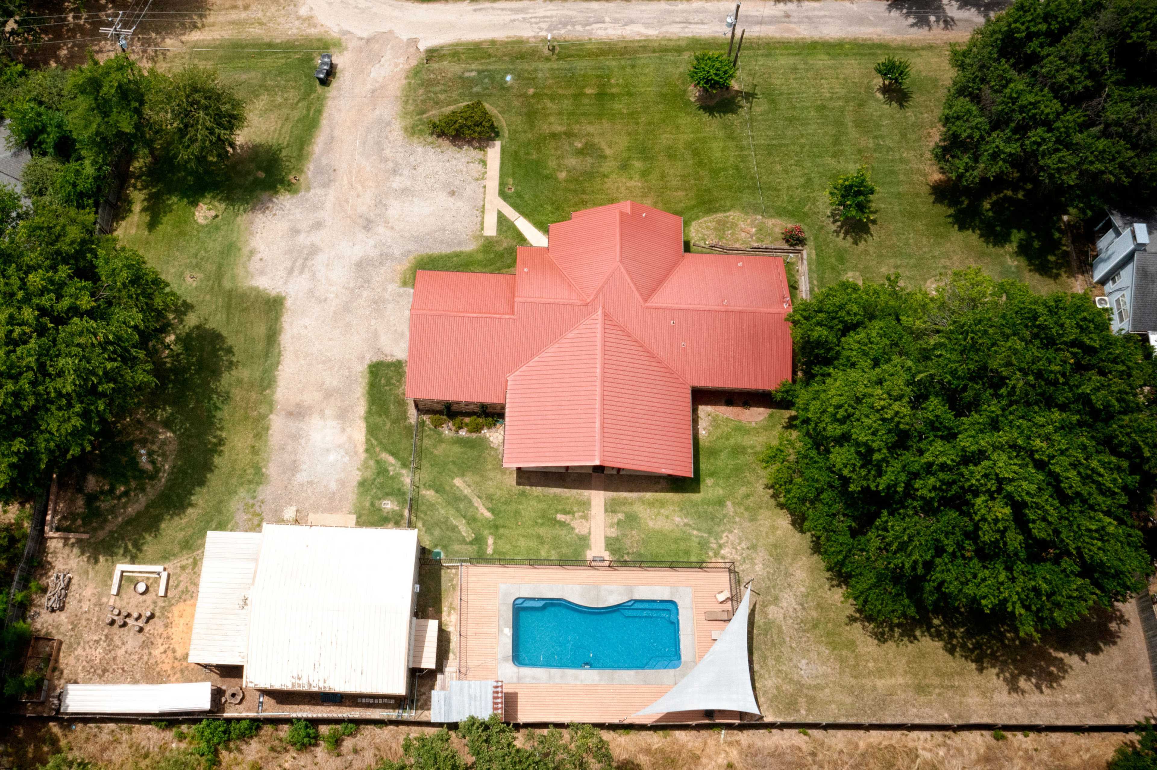 The aerial view shows a large house with a red roof, surrounded by green lawn, a swimming pool, and an adjacent smaller building.