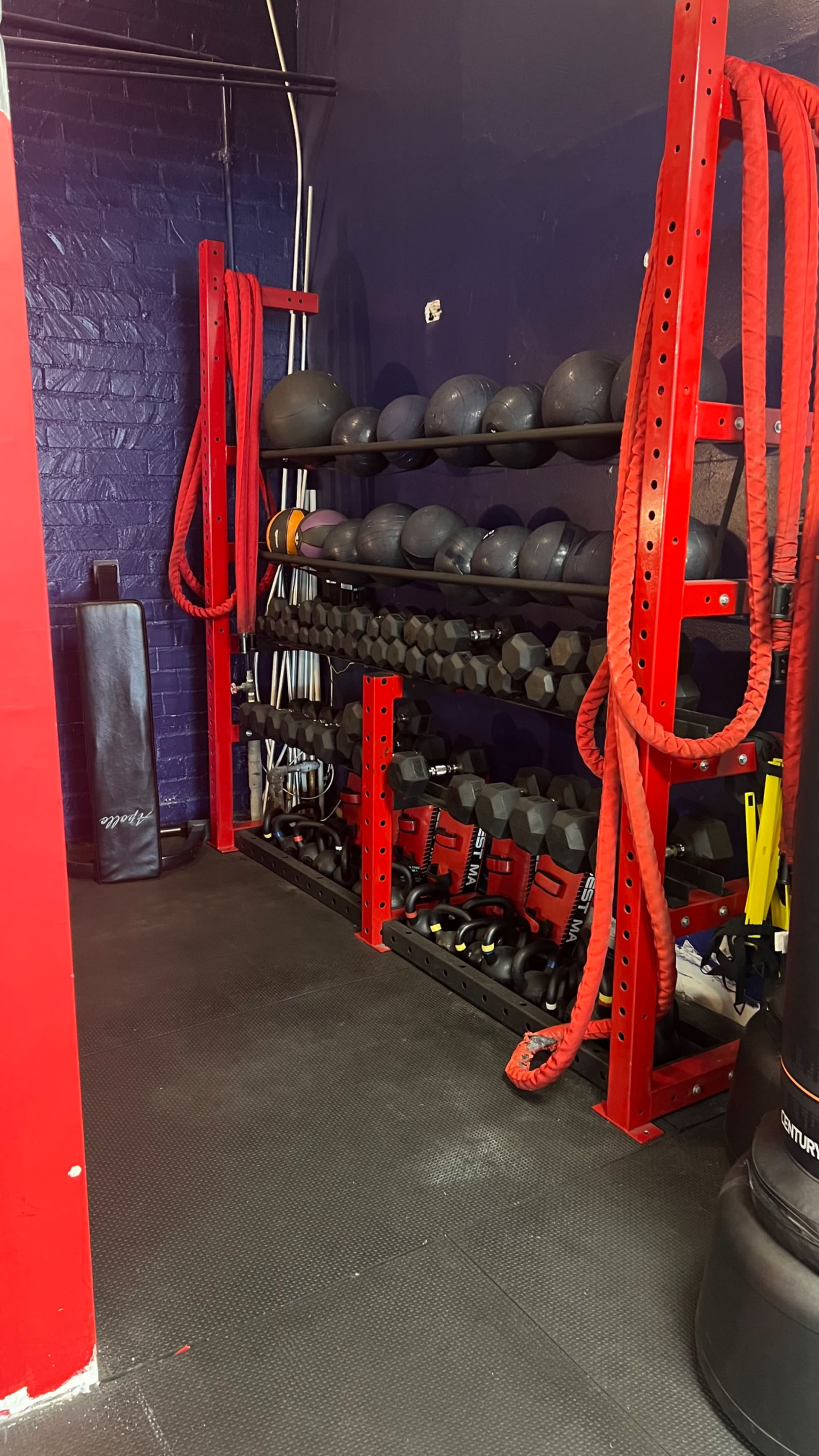 A fitness area with red shelves holding various weights and exercise ropes, alongside a punching bag against a wall.