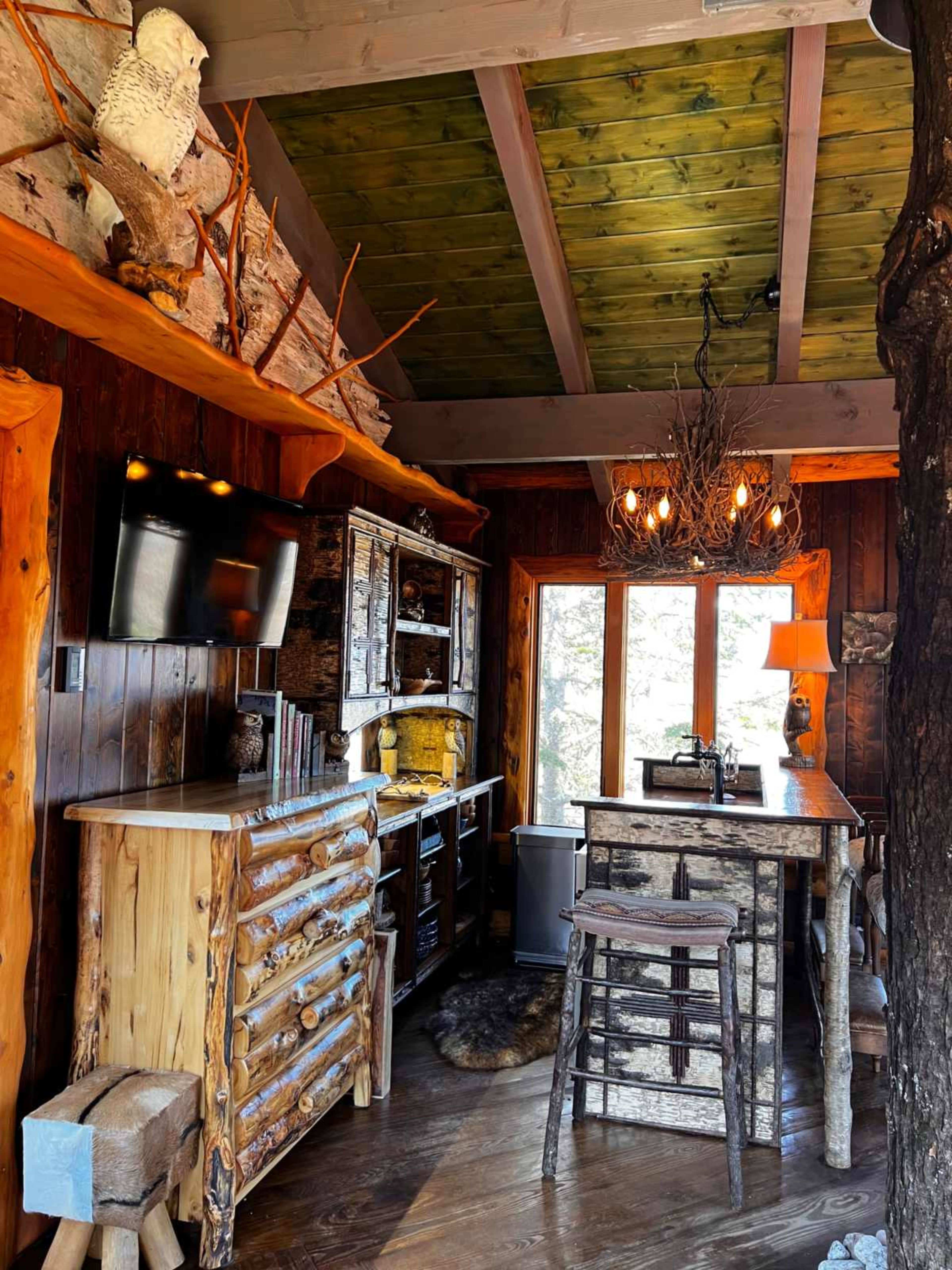 The interior of a rustic cabin features wooden furniture, a chandelier, and a television mounted on the wall.