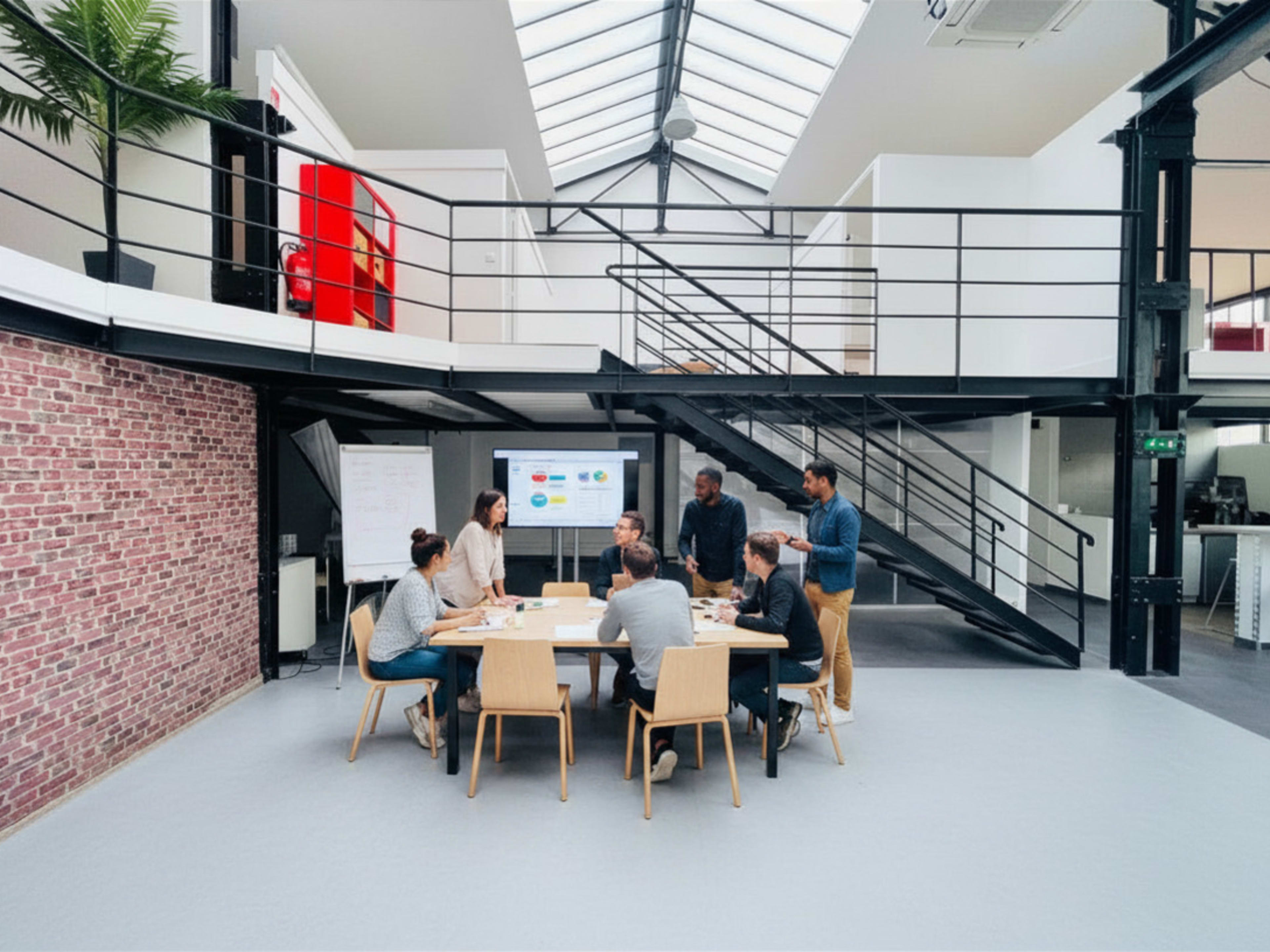 A group of eight people is engaged in a meeting around a large table in a modern office space with a glass roof and exposed brick walls.