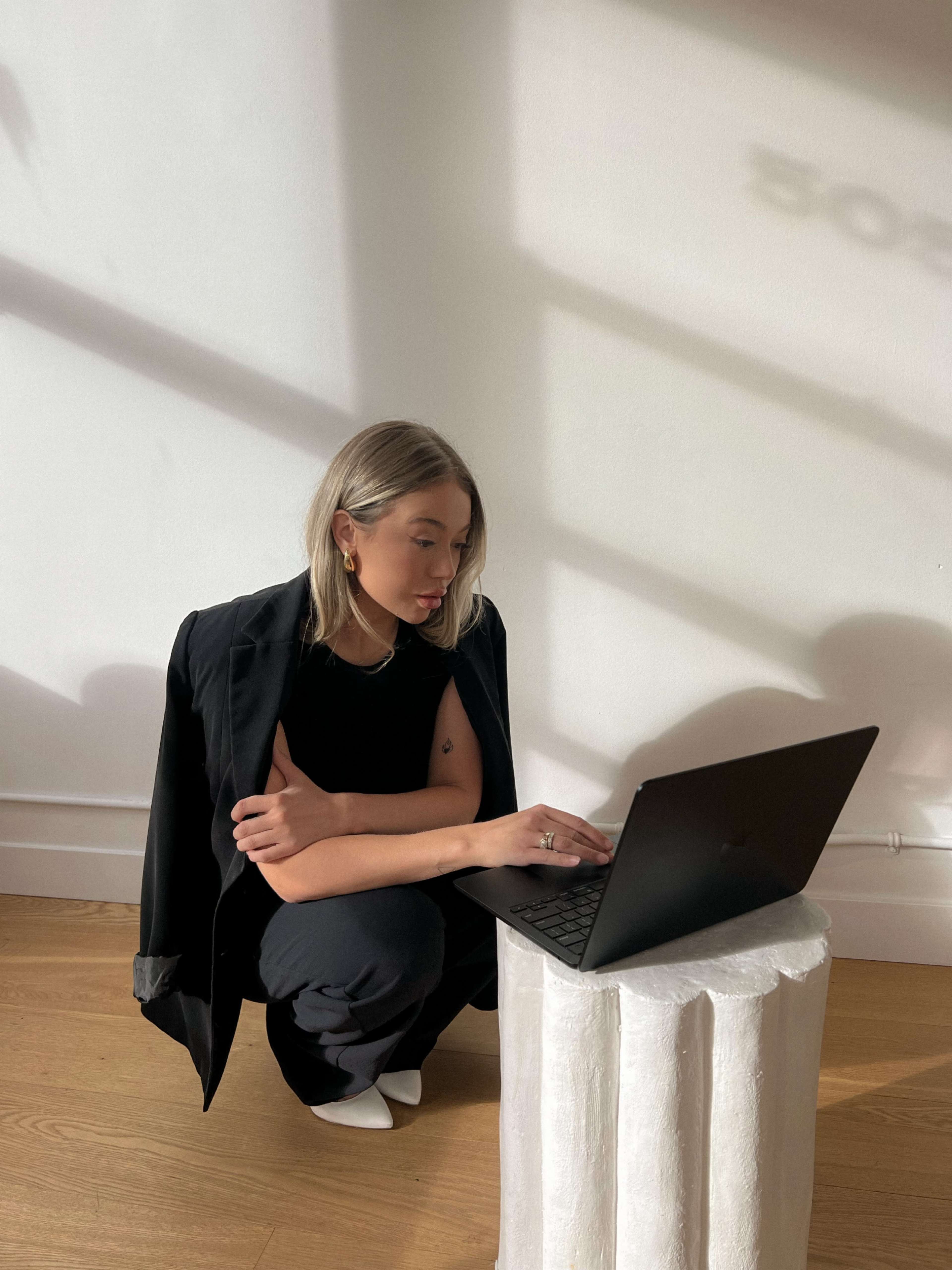 A person crouches beside a white cylindrical table while working on a laptop in a brightly lit room.