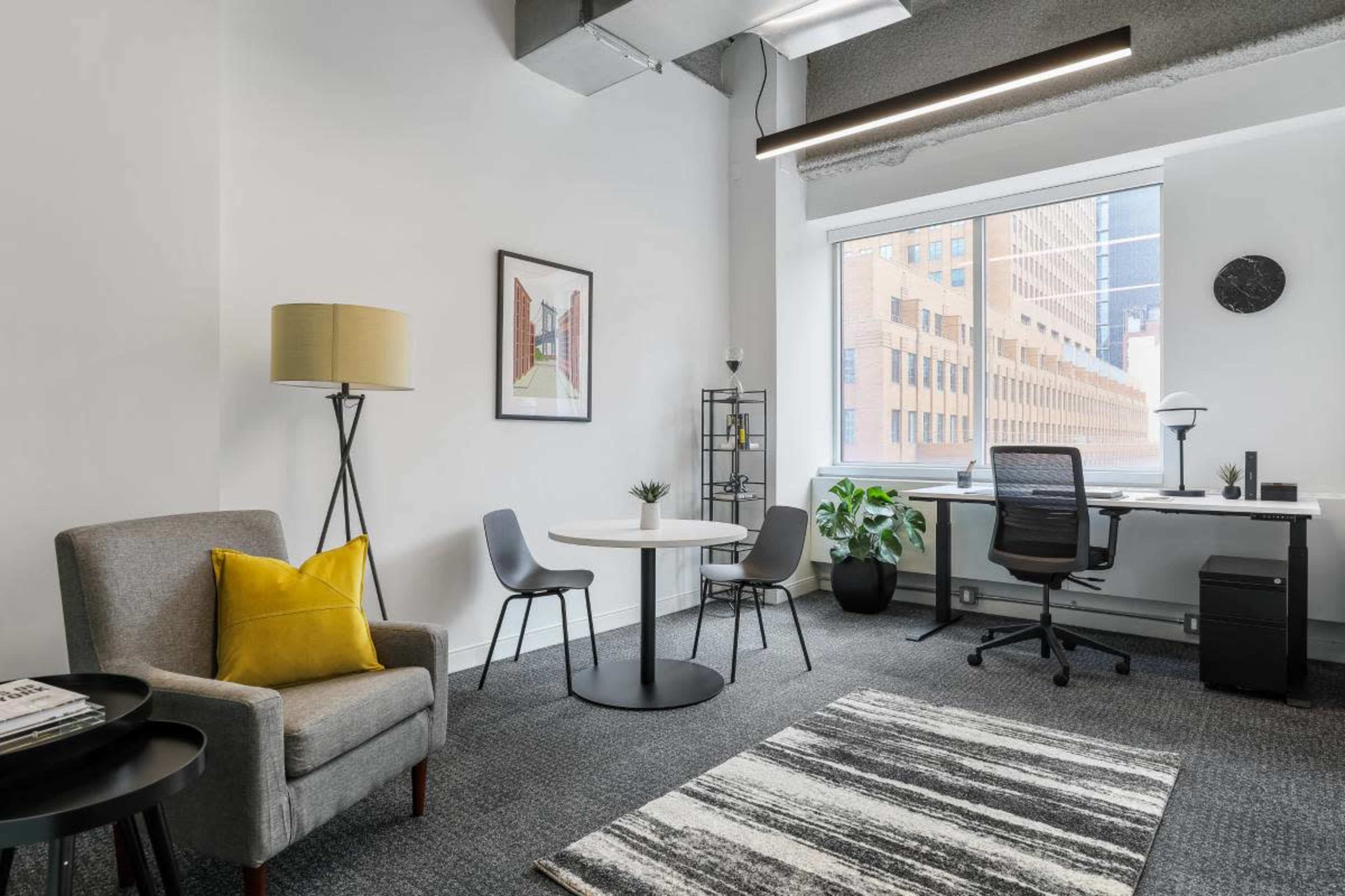 The image shows a modern office space featuring a gray armchair, a small round table, a desk with a chair, and a large window overlooking a cityscape.