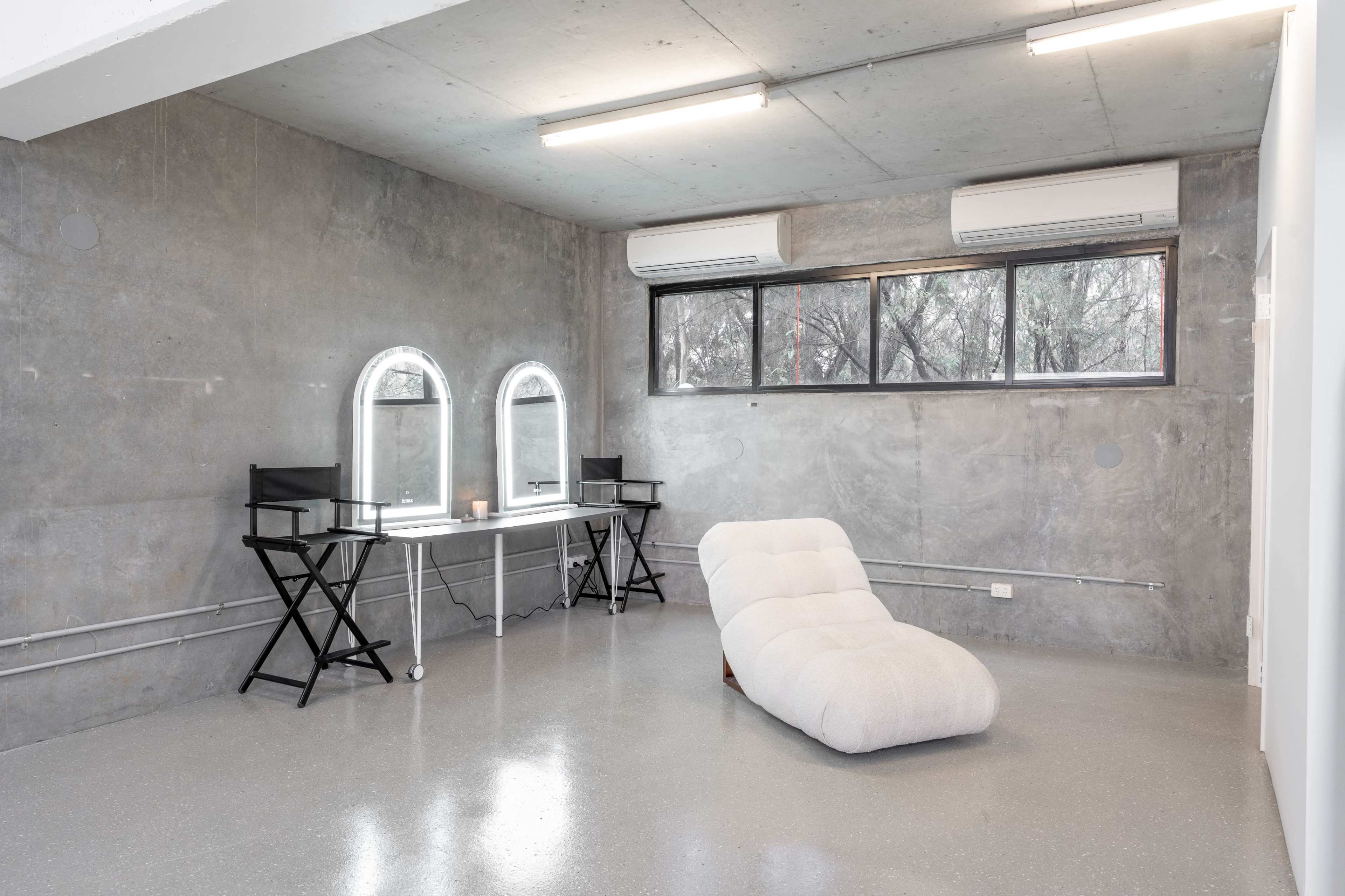 A minimalist room with a concrete wall, a sleek vanity table with two mirrors, a soft chaise lounge, and two folding chairs.