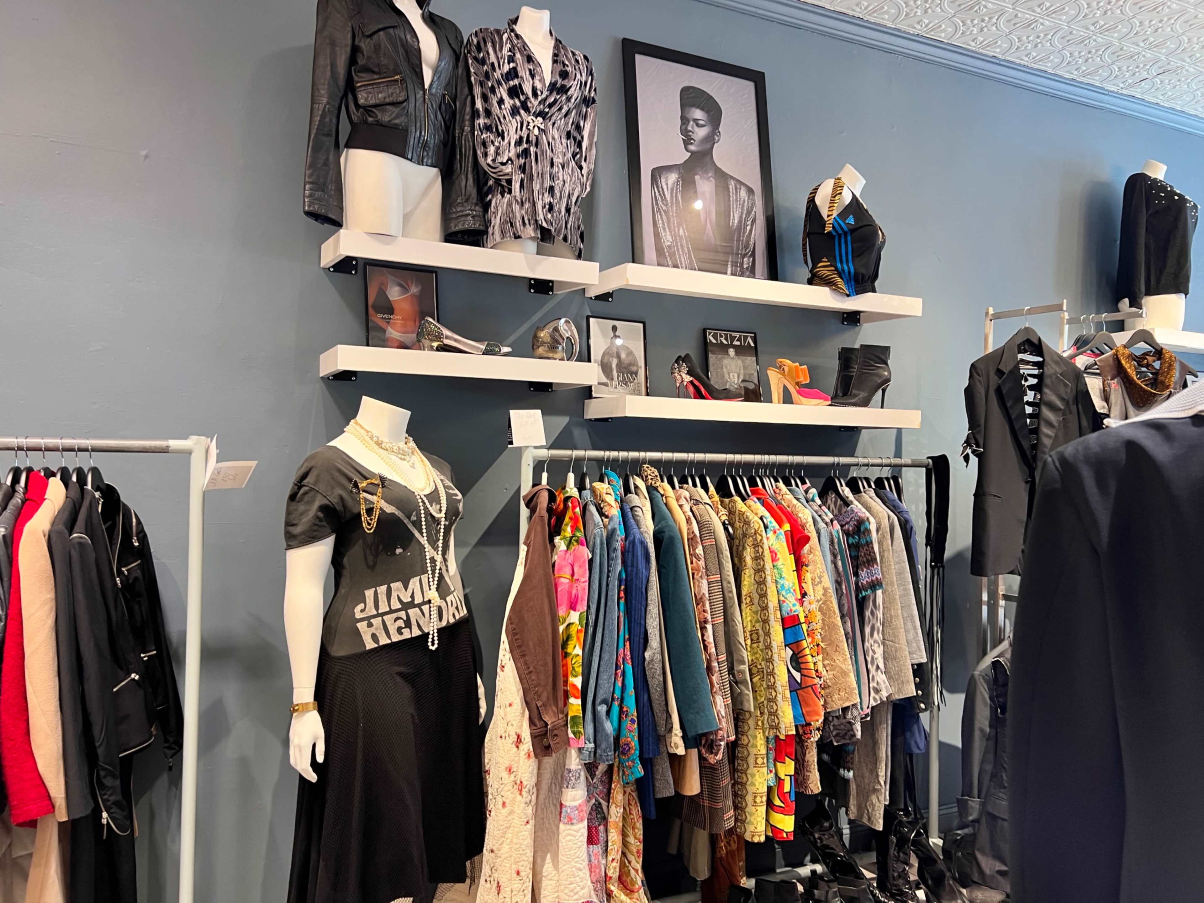 A clothing store displays a variety of apparel on racks and shelves, with items including jackets, dresses, and accessories, alongside a framed portrait on the wall.