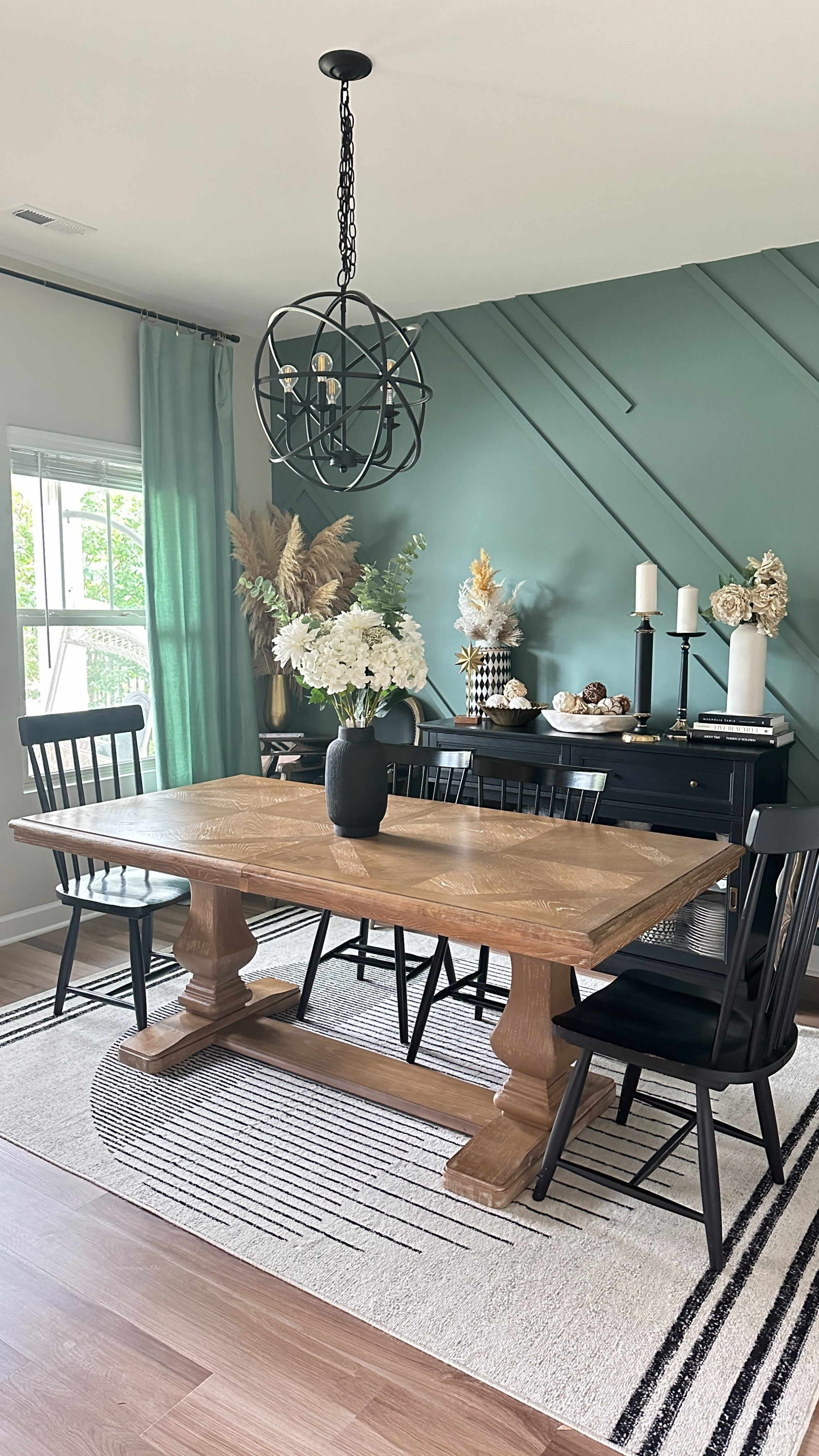 A dining area features a wooden table with black chairs, a vase of flowers, and decorative elements against a green accent wall.