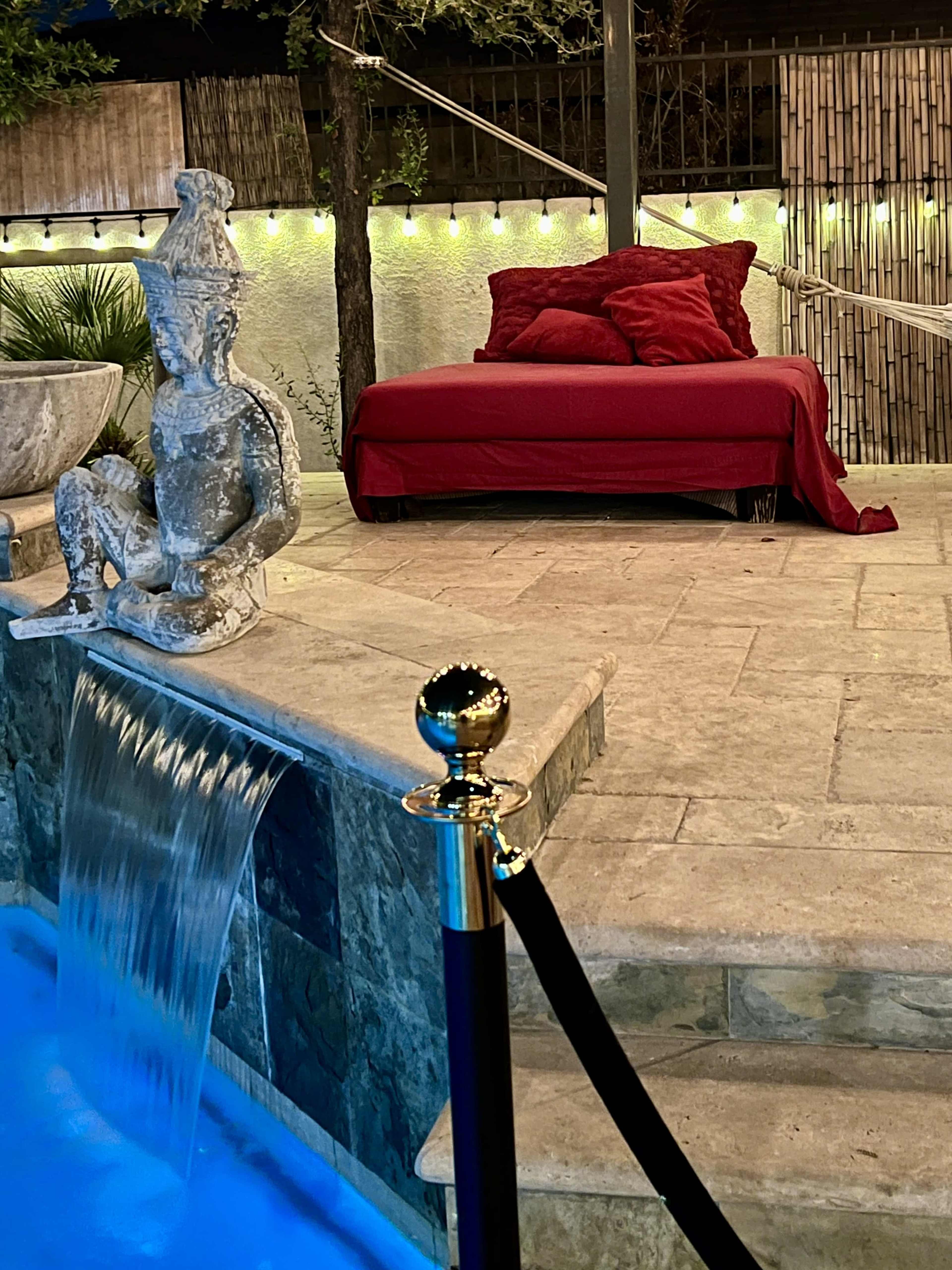 A small stone waterfall flows into a pool beside a red-covered daybed and a statue seated nearby, with string lights illuminating the area.