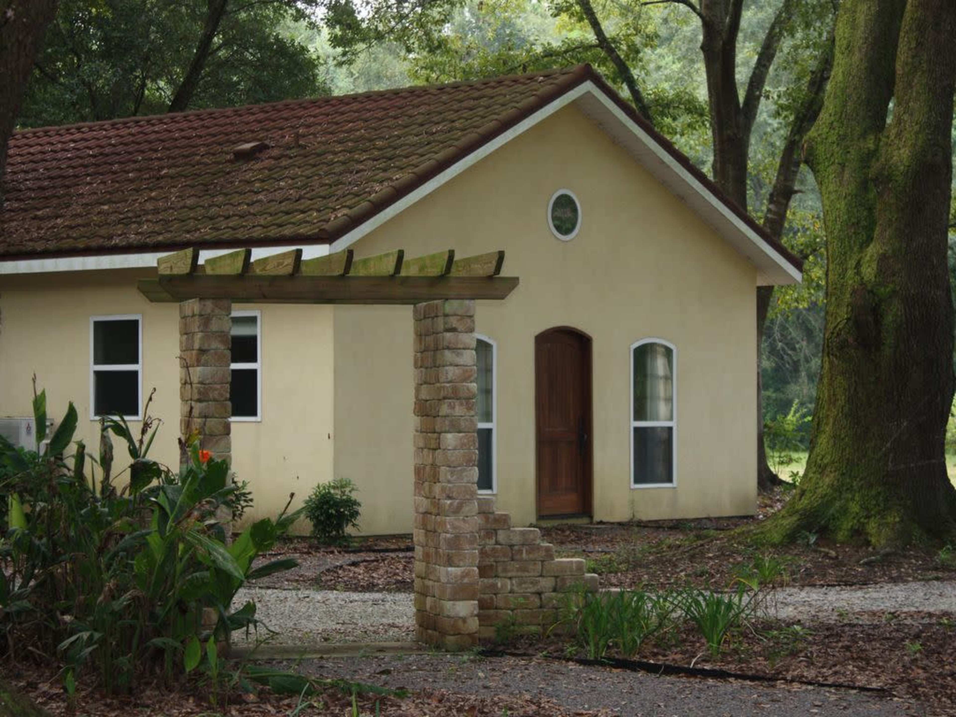 A small house with a brown tiled roof is situated behind a stone archway, surrounded by trees and greenery.