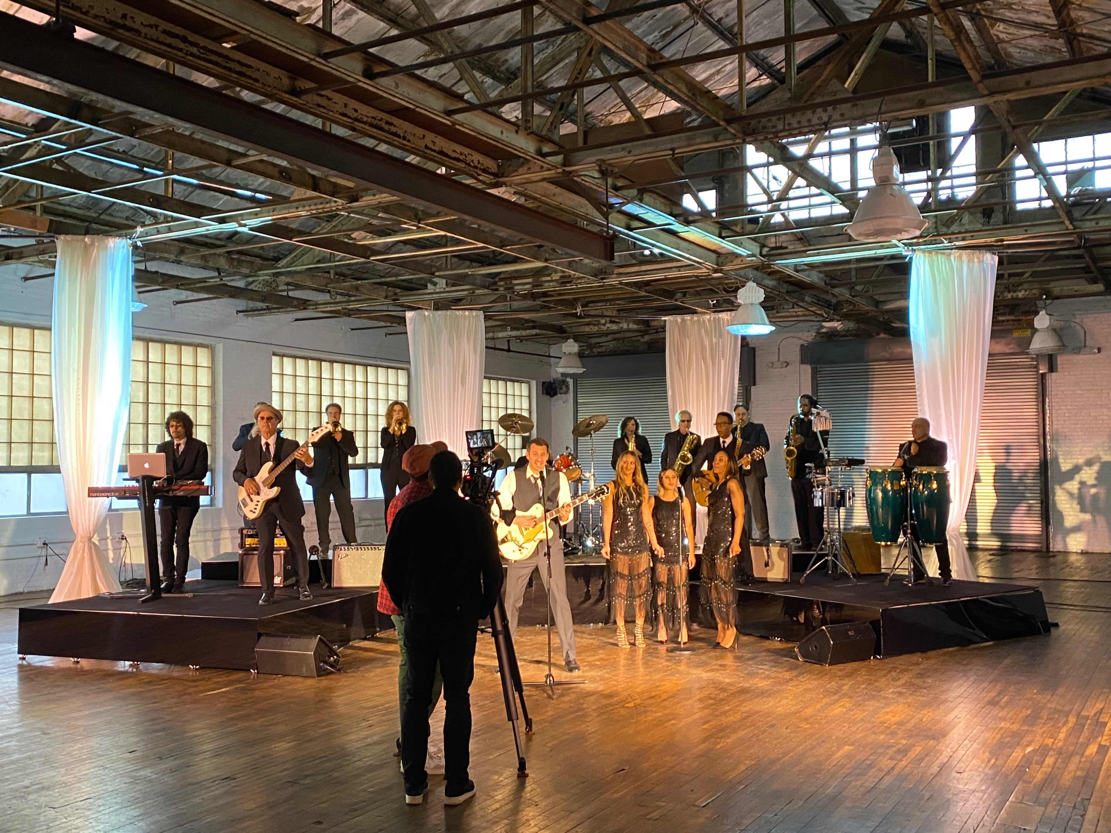 A live band performs on a stage in a large, industrial space with various instruments and musicians, while a camera crew records the performance.
